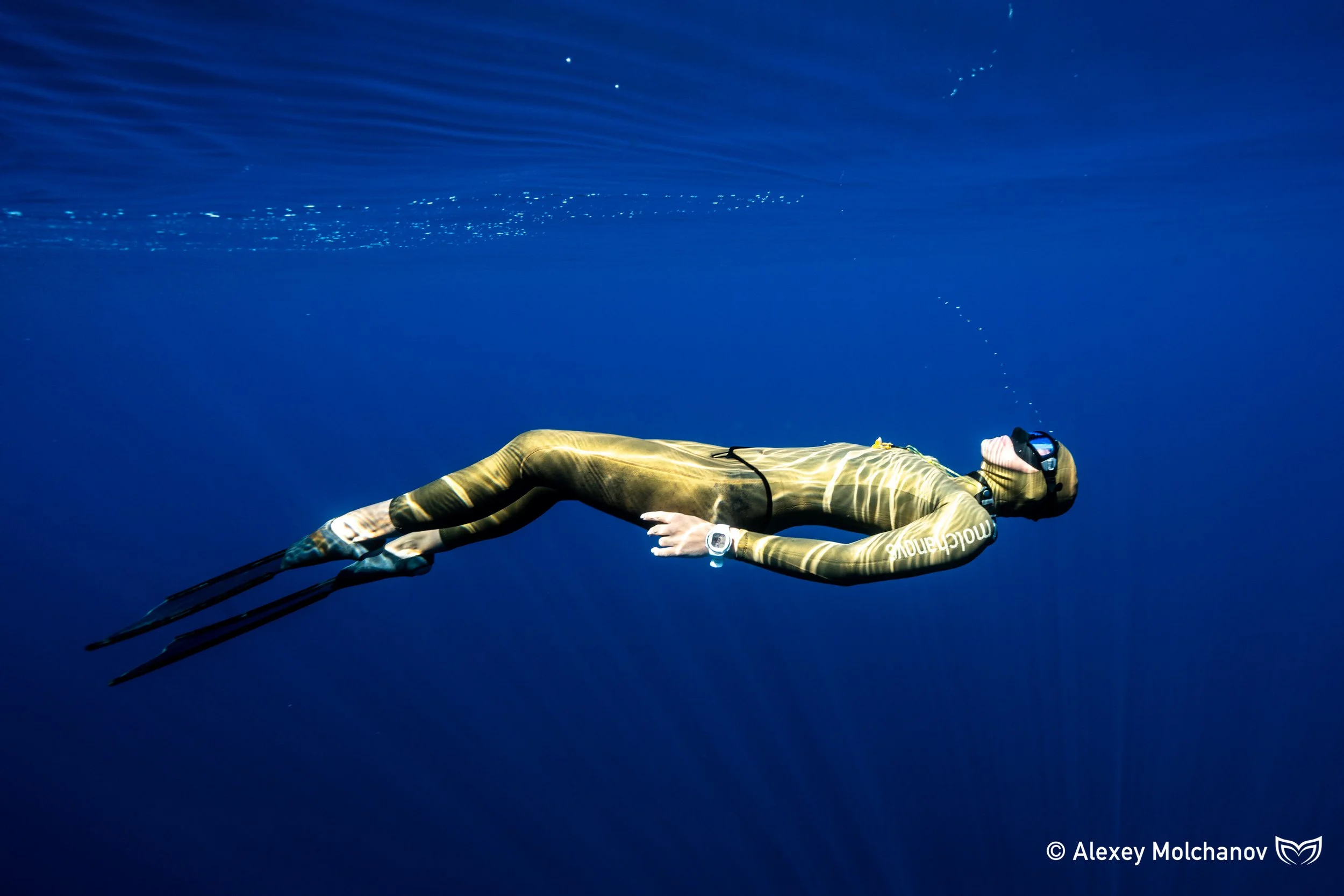A woman in a wetsuit and snorkeling gear floating face down in the deep blue ocean with a trail of bubbles ascending to the surface.