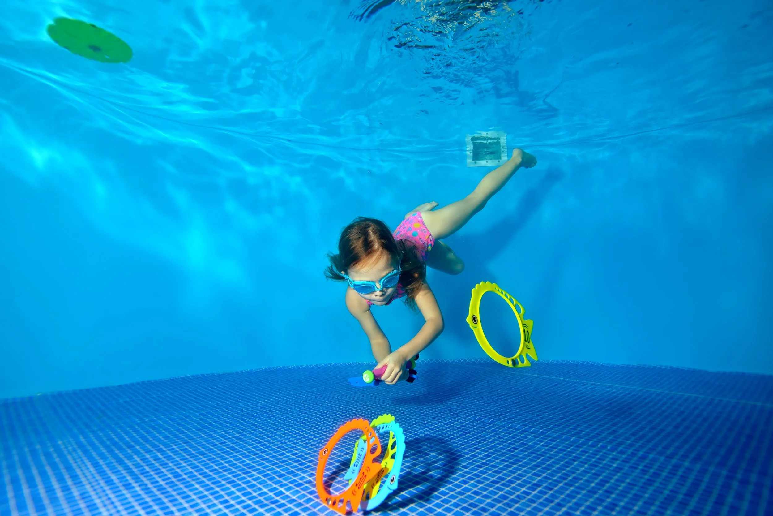 A young girl swimming underwater in a swimming pool, holding colorful underwater toys and wearing goggles.