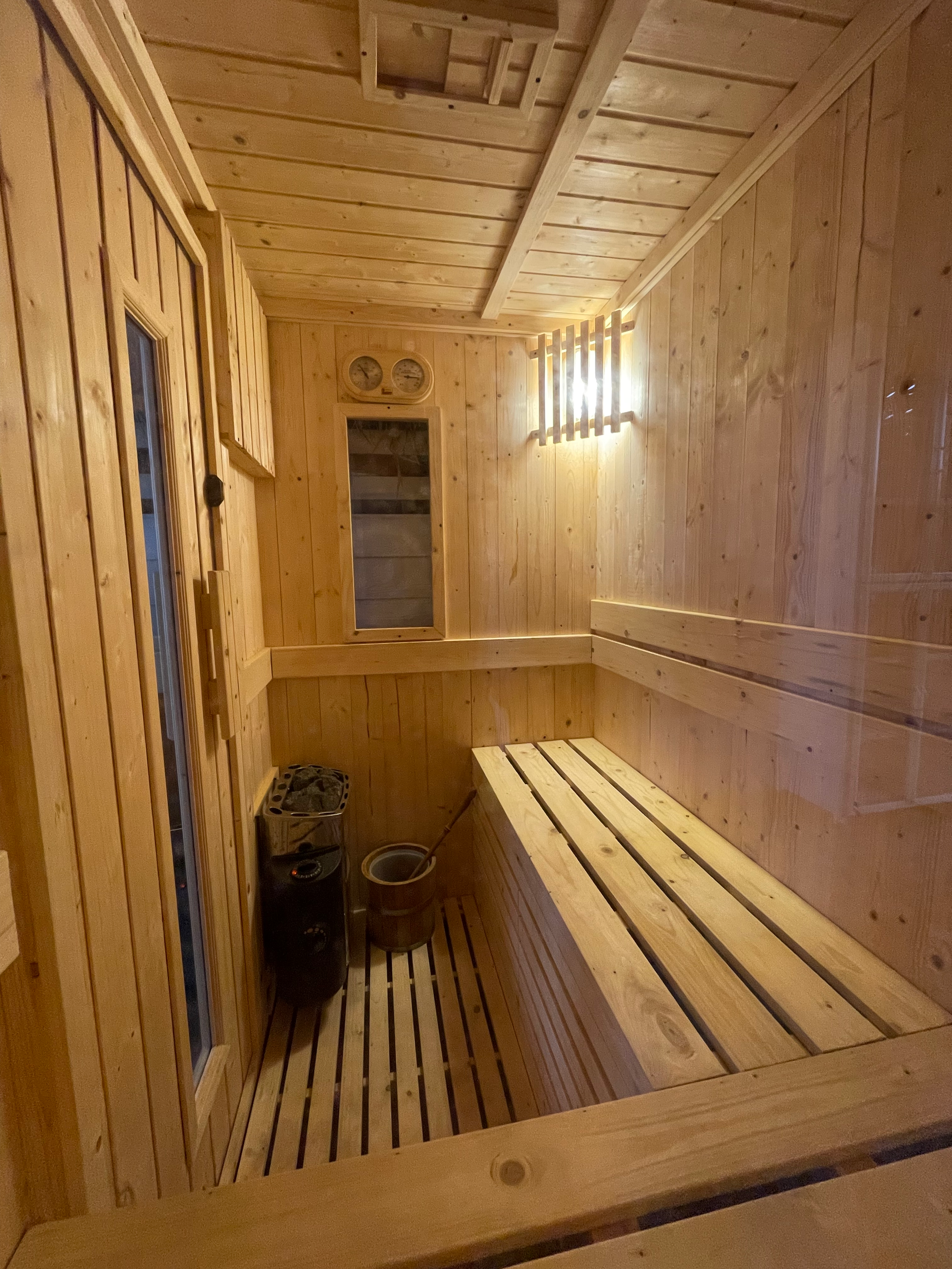 The image shows the interior of a small wooden sauna with wood-paneled walls, benches, a heater, and a thermometer and hygrometer mounted on the back wall.
