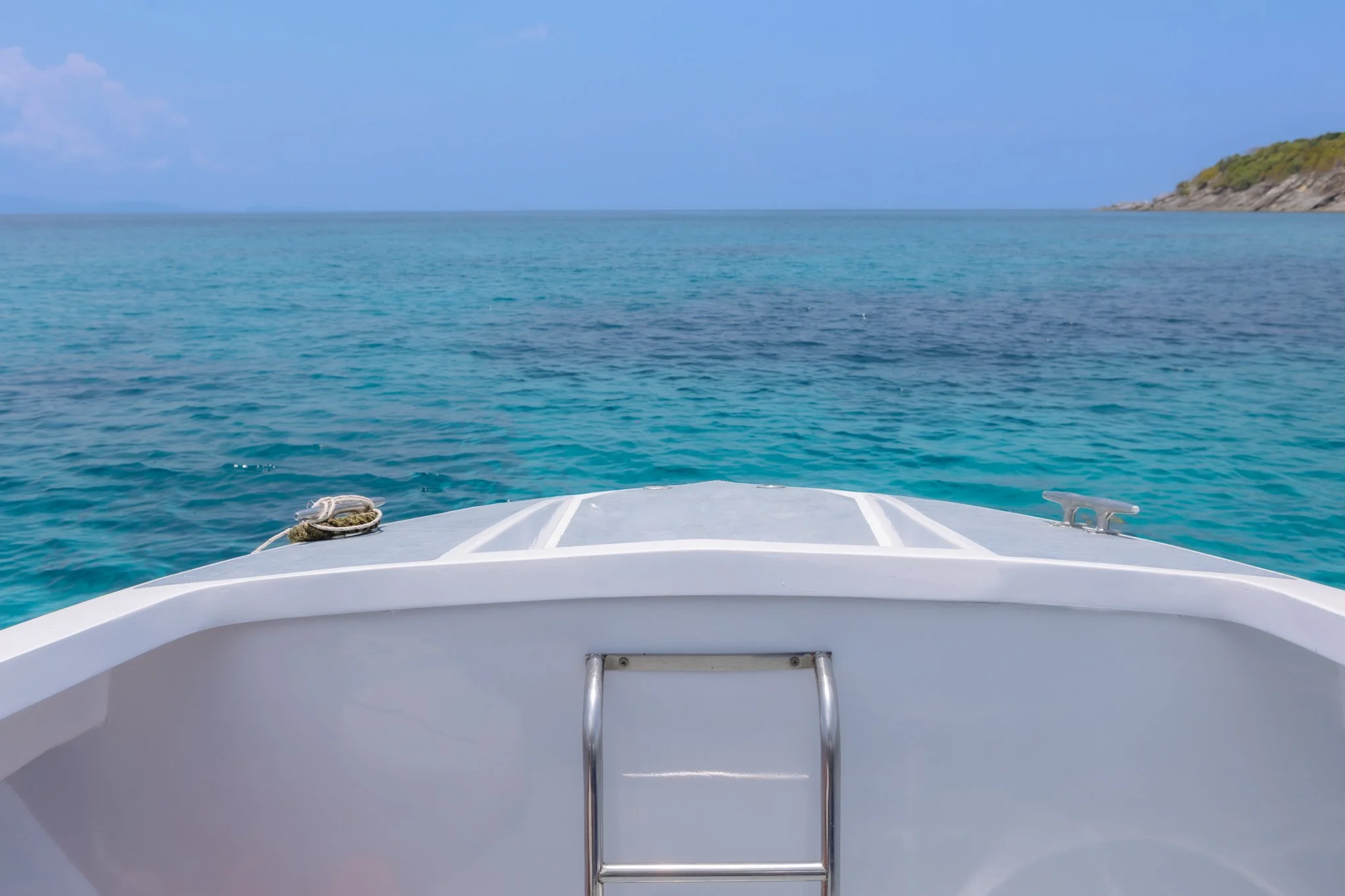 View from the bow of a boat looking out over clear blue water towards a distant shoreline with green foliage under a partly cloudy sky.