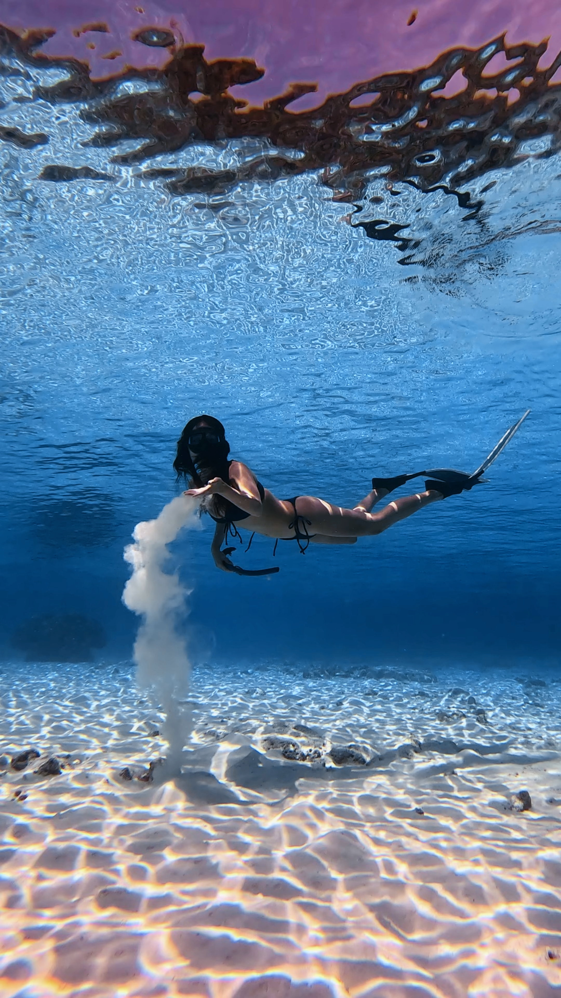 A woman in a bikini and snorkeling gear underwater, holding a snorkel tube in her right hand, with water and rocks visible on the ocean floor.