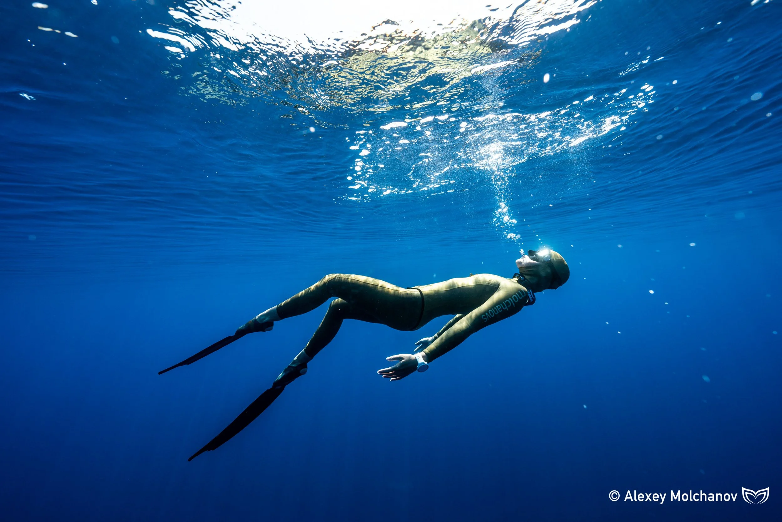 A woman in a blue wetsuit with goggles holds a long pole while scuba diving underwater.