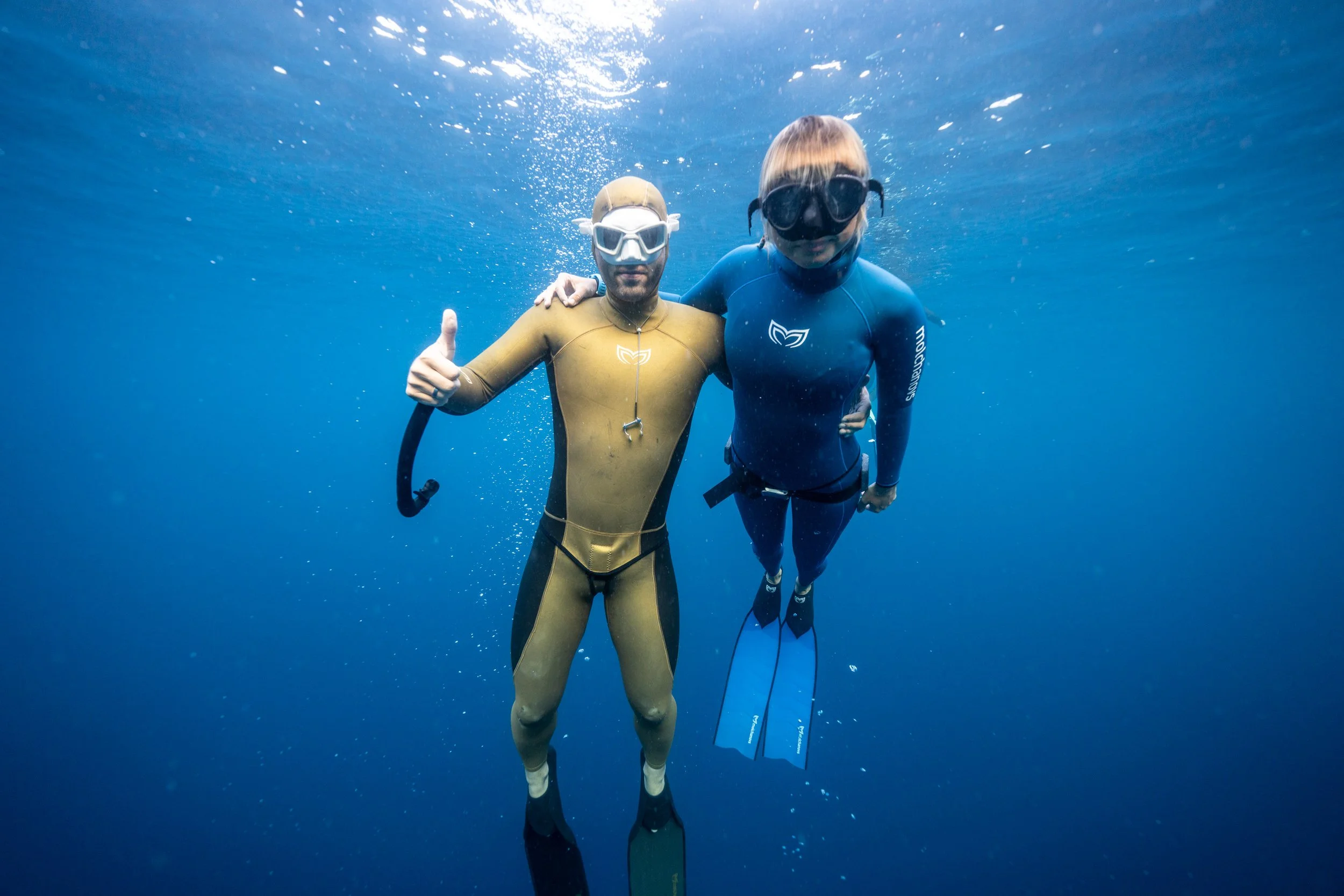 Two people in wetsuits and snorkeling gear underwater, one giving a thumbs-up