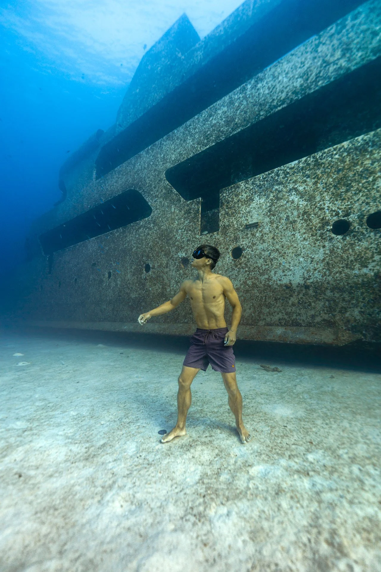 A man with freediving mask and swim trunks standing underwater next to a large, rusted shipwreck.