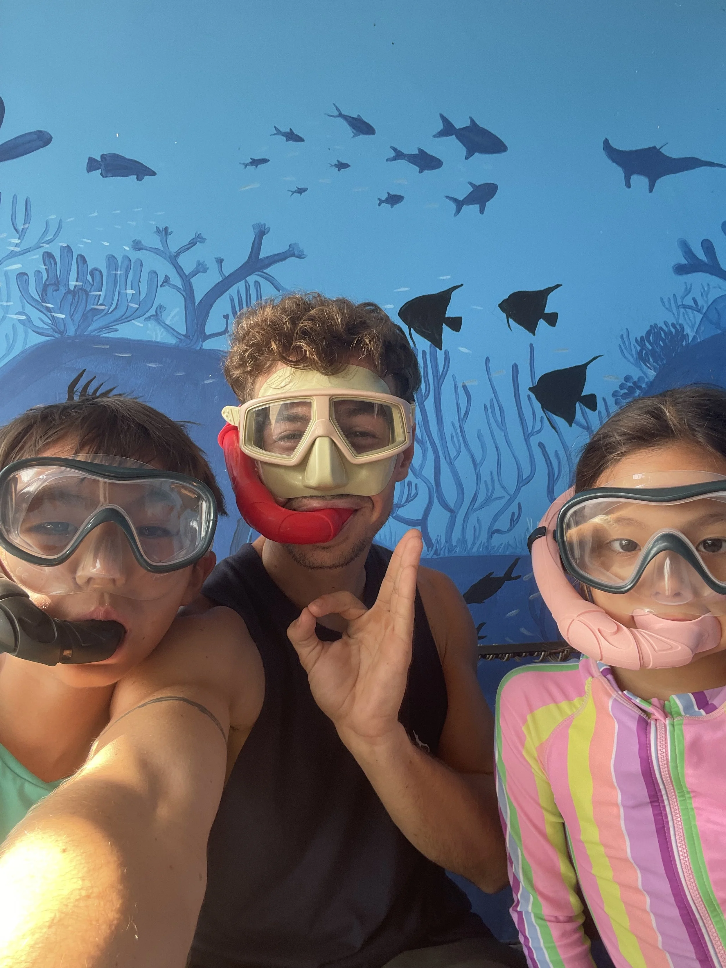 Three people in snorkeling gear taking a selfie underwater with a blue backdrop of fish and coral.