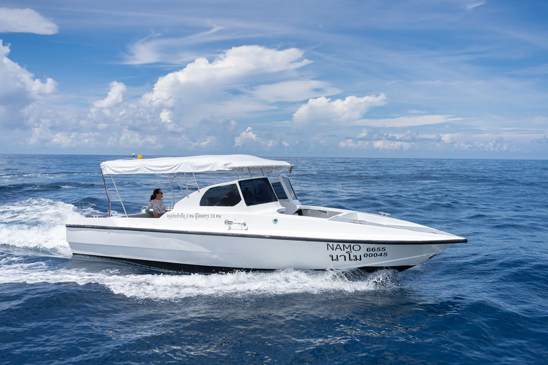 A white motorboat named NAMO traveling on the open ocean with a woman seated inside, under a partly cloudy sky.