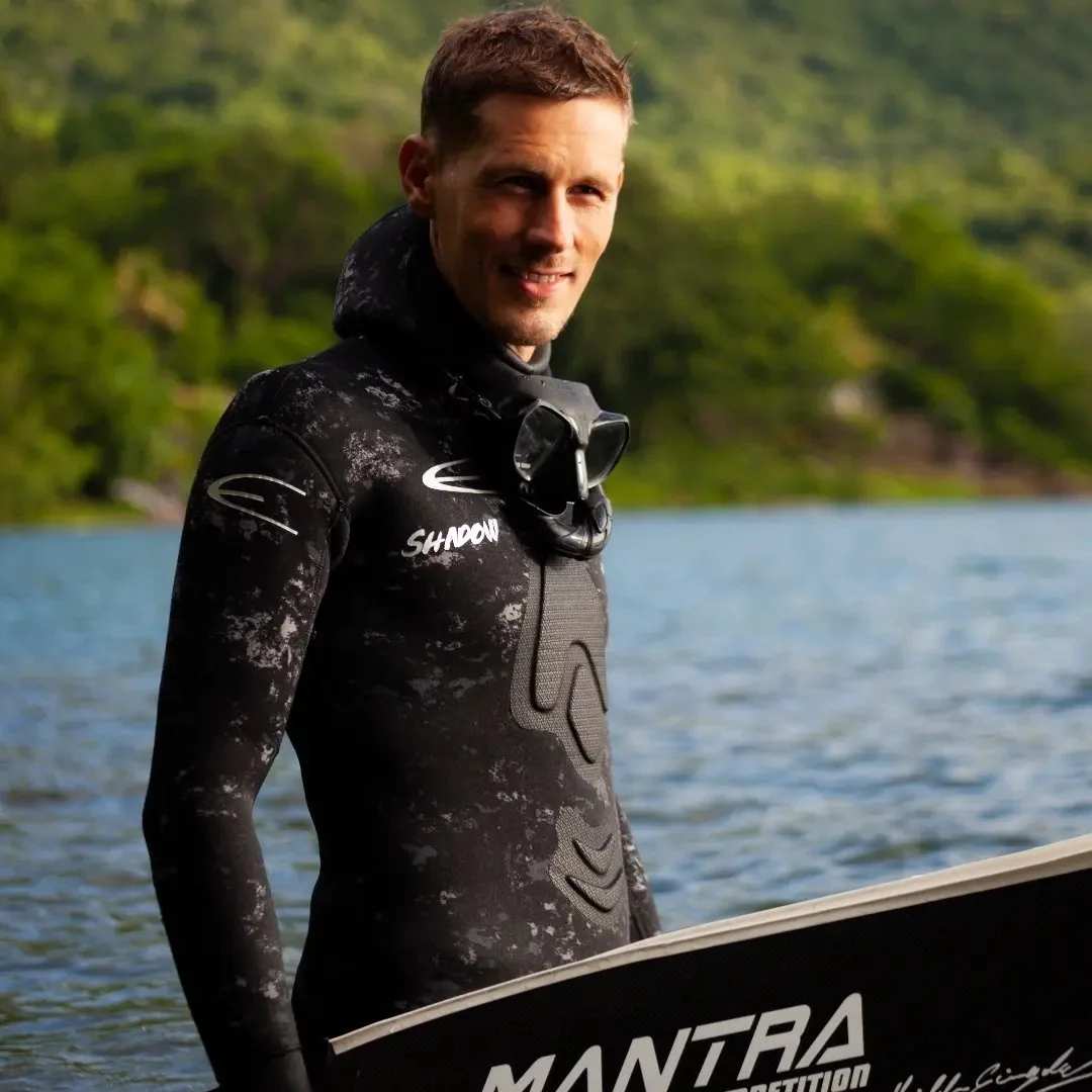 A man in a wetsuit holding a wakeboard near a body of water with green trees in the background.