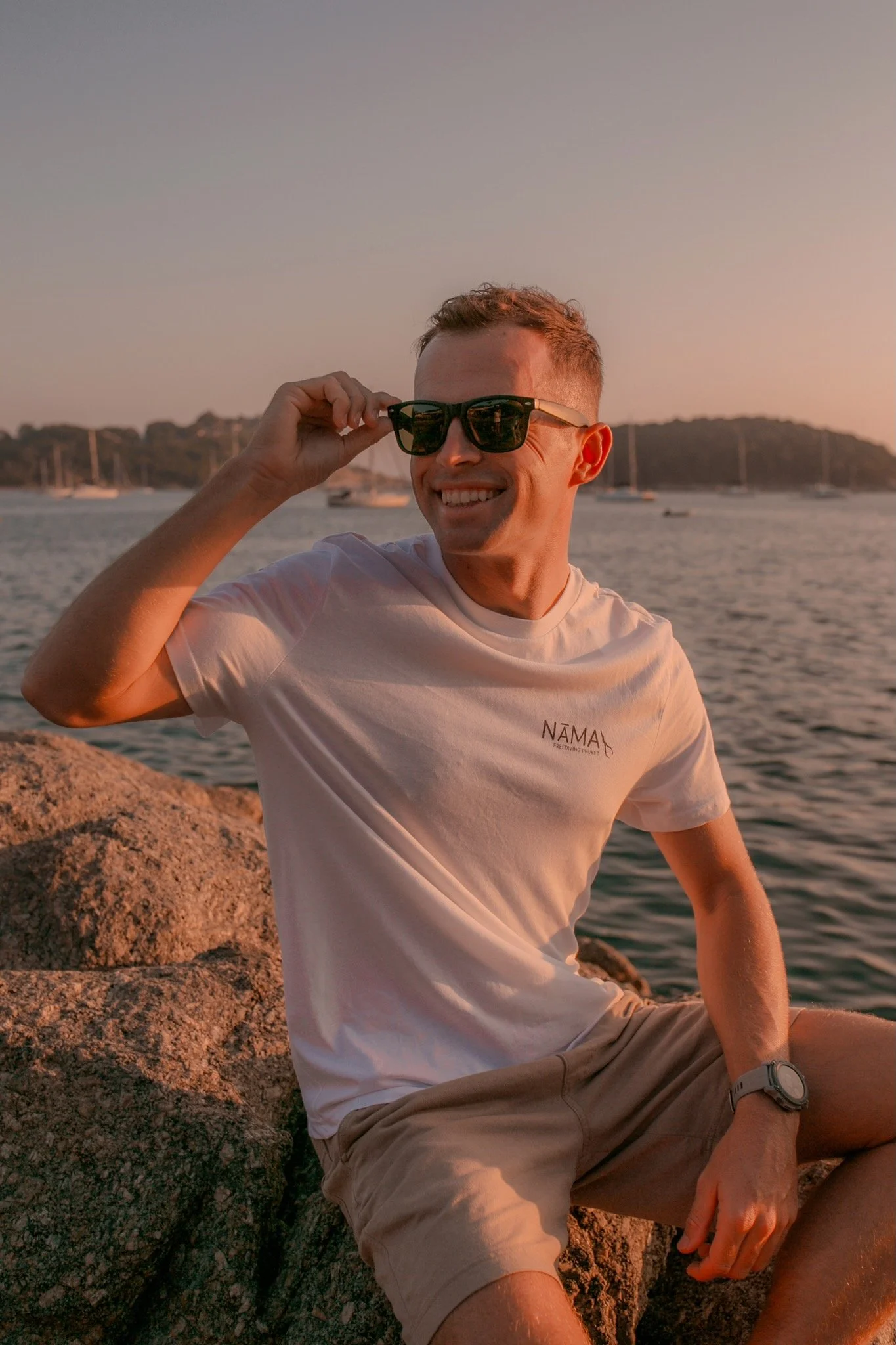 A young man with short hair and sunglasses smiling, sitting on a rock near a body of water with boats and a hilly shoreline in the background during sunset.