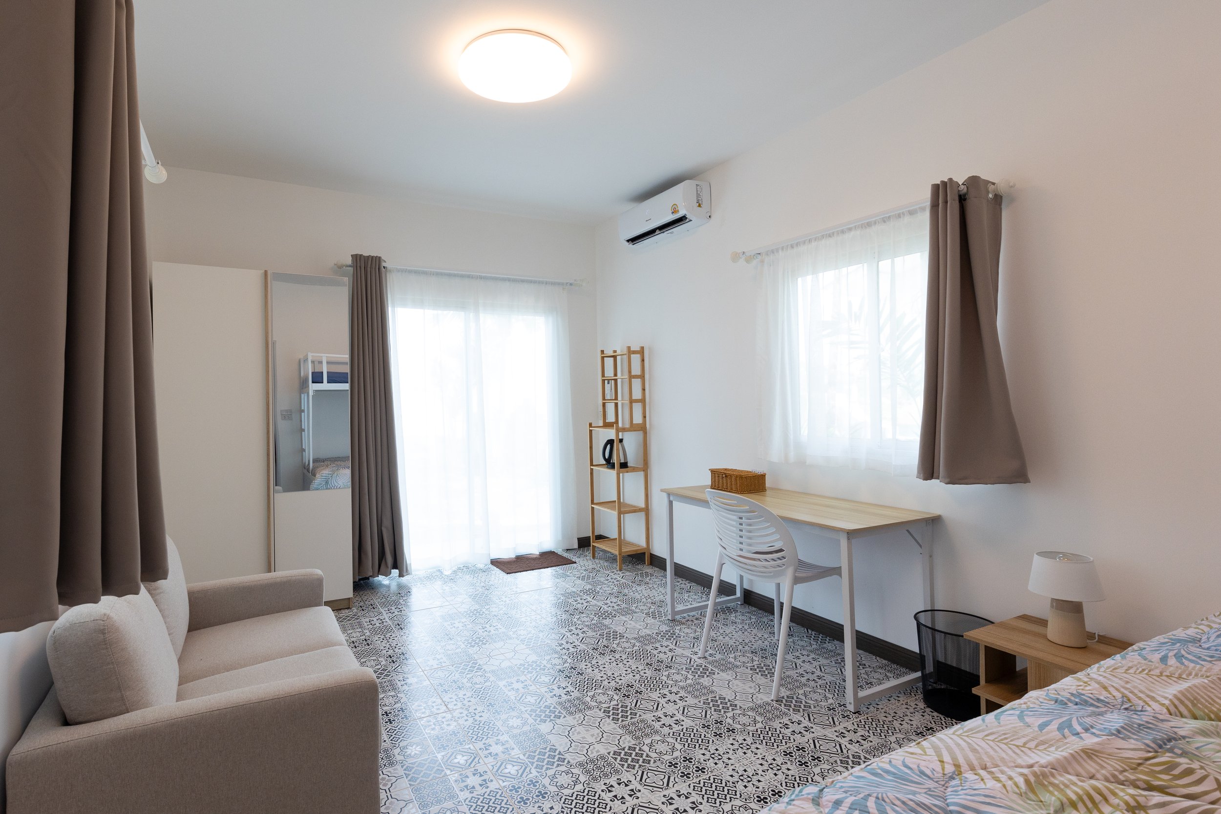 Interior of a bright, minimalist living space with large windows, curtains, a white sofa, a wooden desk with a white chair, a wicker basket, a small lamp, a mirror, and patterned tile flooring.