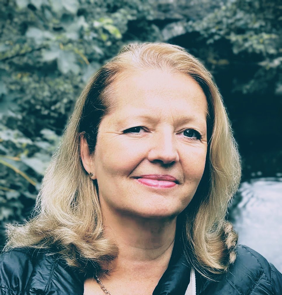 A middle-aged woman with blonde hair smiling outdoors with greenery and a river in the background.