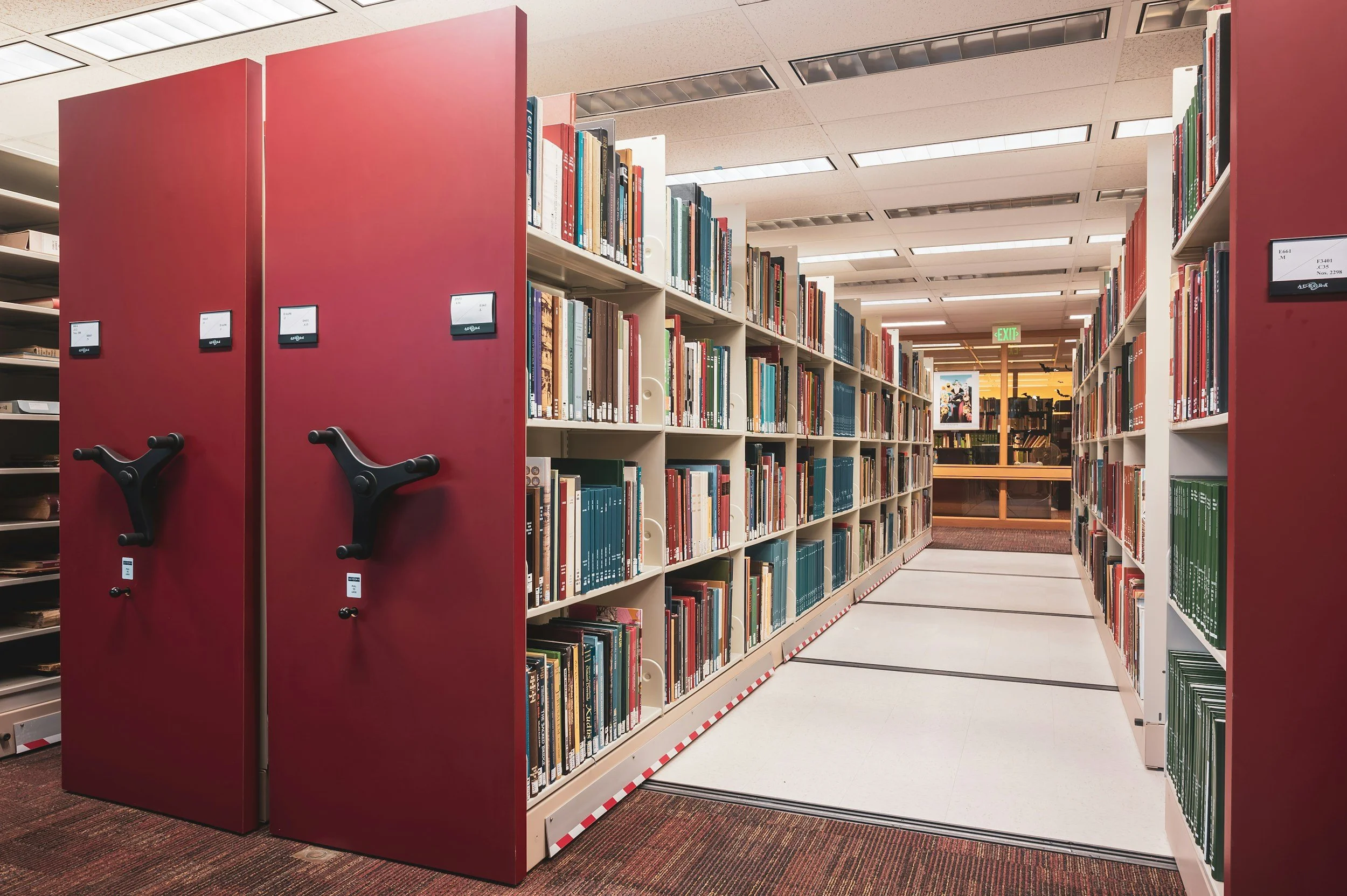 Bookshelves in a library with a red aisle divider and an exit sign in the background.