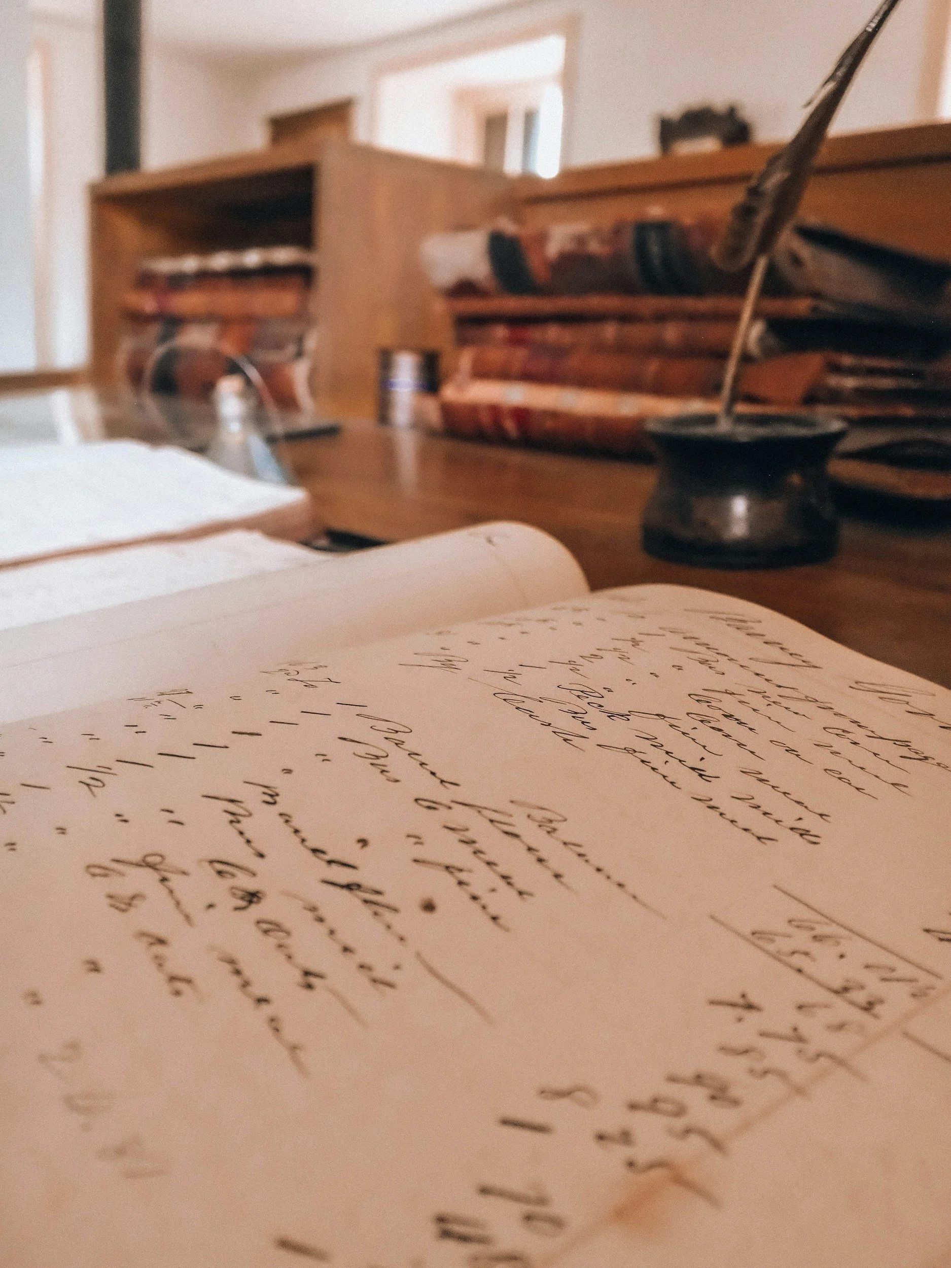 Close-up of handwritten notes on a piece of paper, with a wooden desk, a stack of old books, and a quill pen in inkpot in the background.