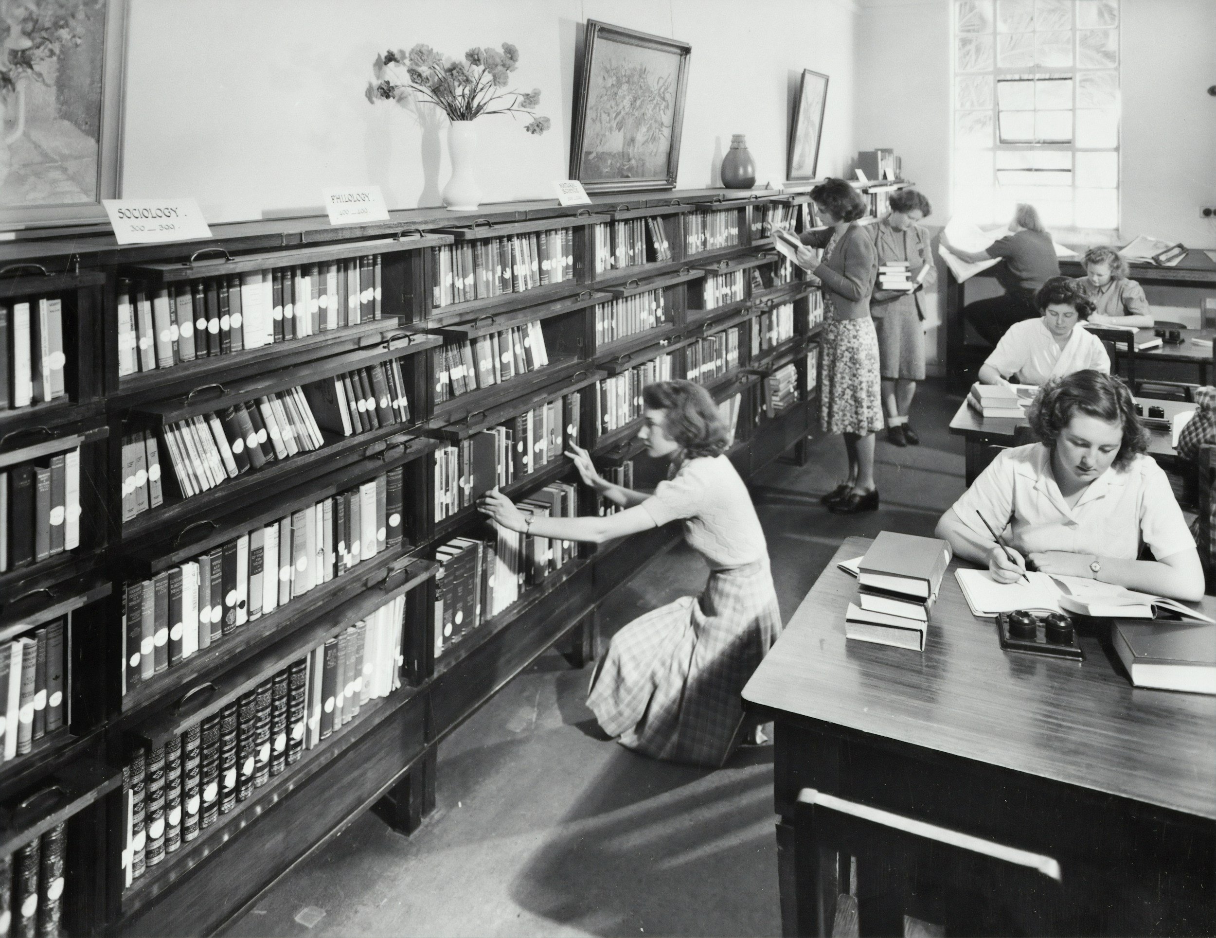 A black and white photo of a library or bookstore with women and girls browsing books on long wooden shelves and tables, with a large window in the background.