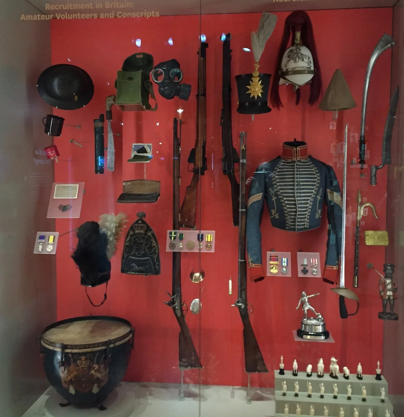 Display of historical military artifacts including rifles, medals, uniforms, swords, helmets, and chess pieces in a museum exhibit.