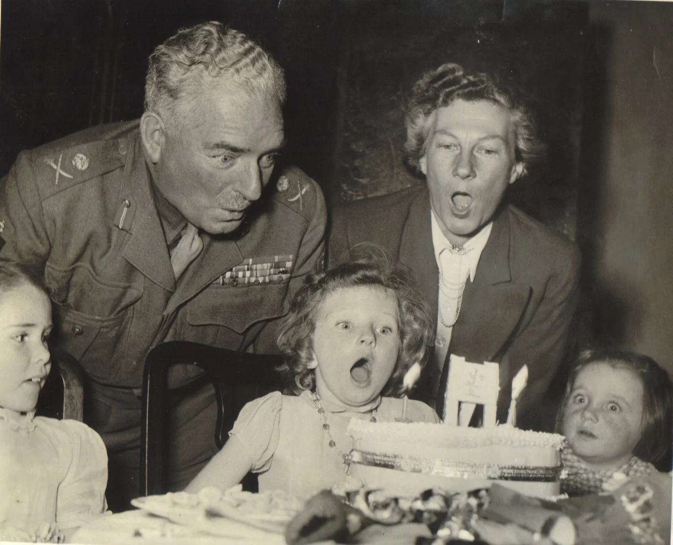 Black and white photo of children celebrating a birthday with a cake, with two adults, one in military uniform, looking surprised or excited.
