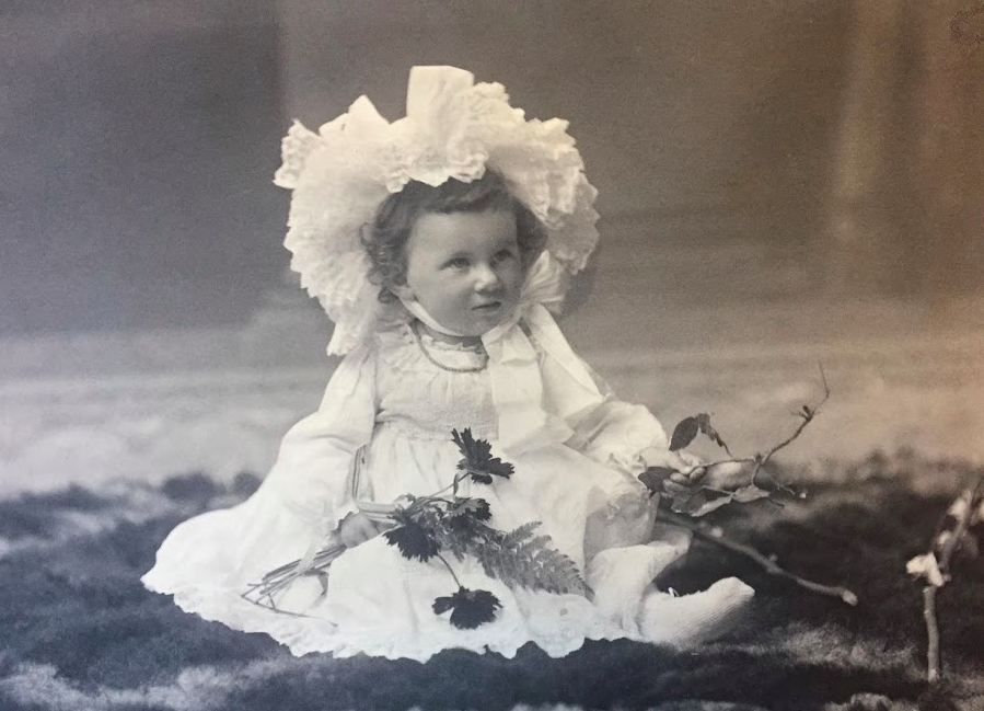 A young girl wearing a large lace bonnet and a long dress sitting on the ground. She is holding a small branch with leaves.
