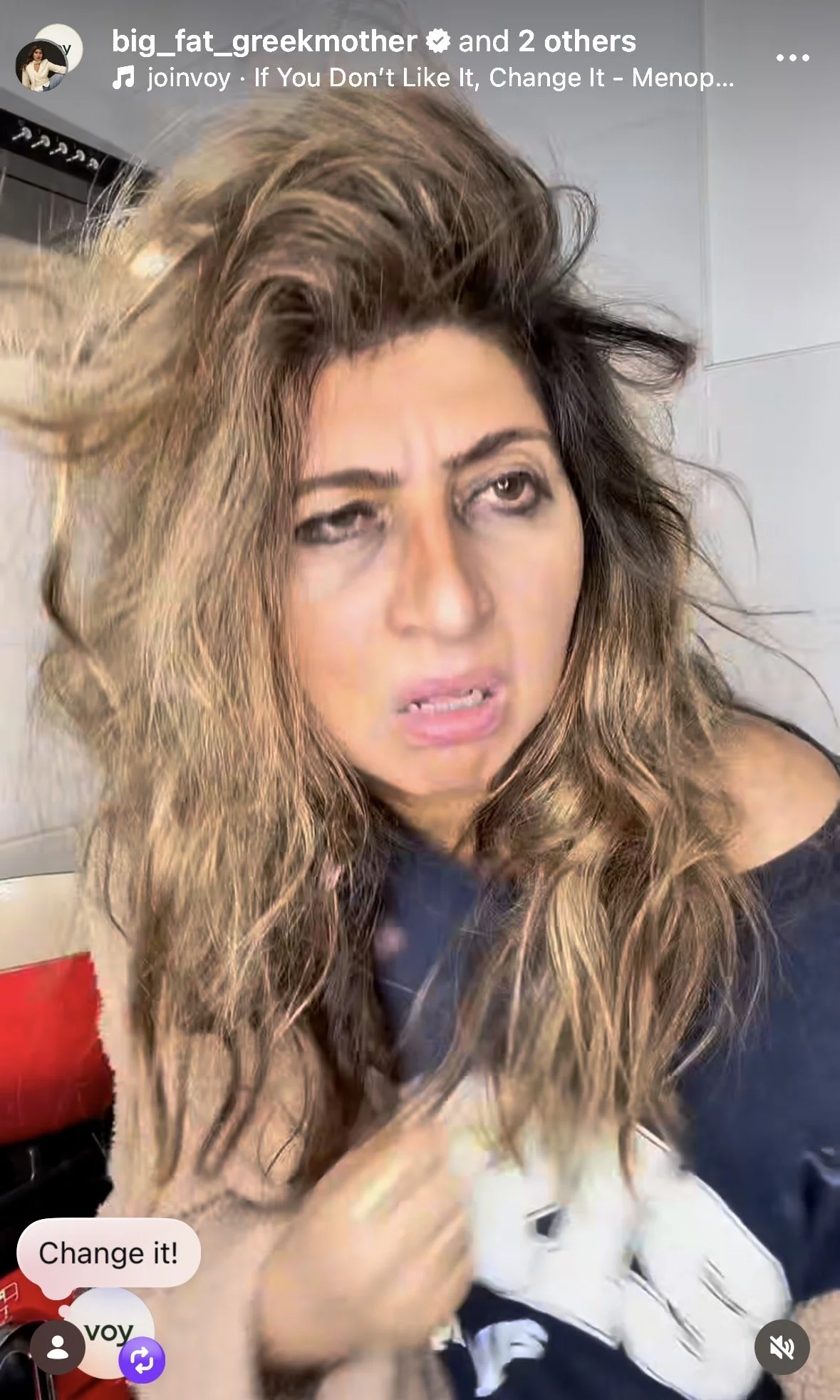 A woman with long, curly, light brown hair and a black off-shoulder top appears to be slightly confused or annoyed. She is situated indoors, possibly in a kitchen or dining area, with tiled walls in the background.