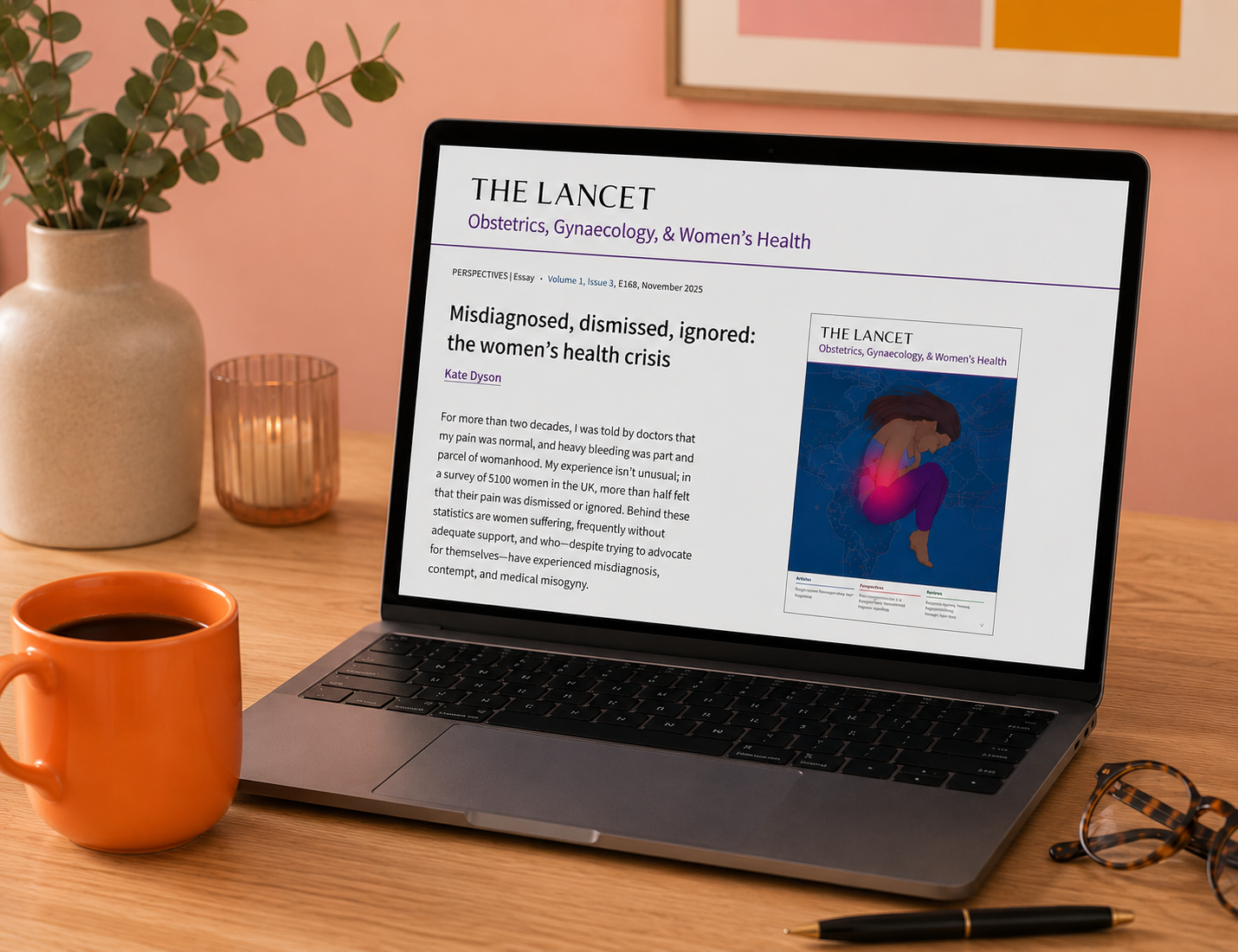A laptop on a wooden desk displays an article from The Lancet about women's health, with a pink background wall and a framed art print behind it. The desk holds an orange coffee mug, a pair of glasses, and a black pen. To the left, a beige vase with a plant and a small candle holder are visible.