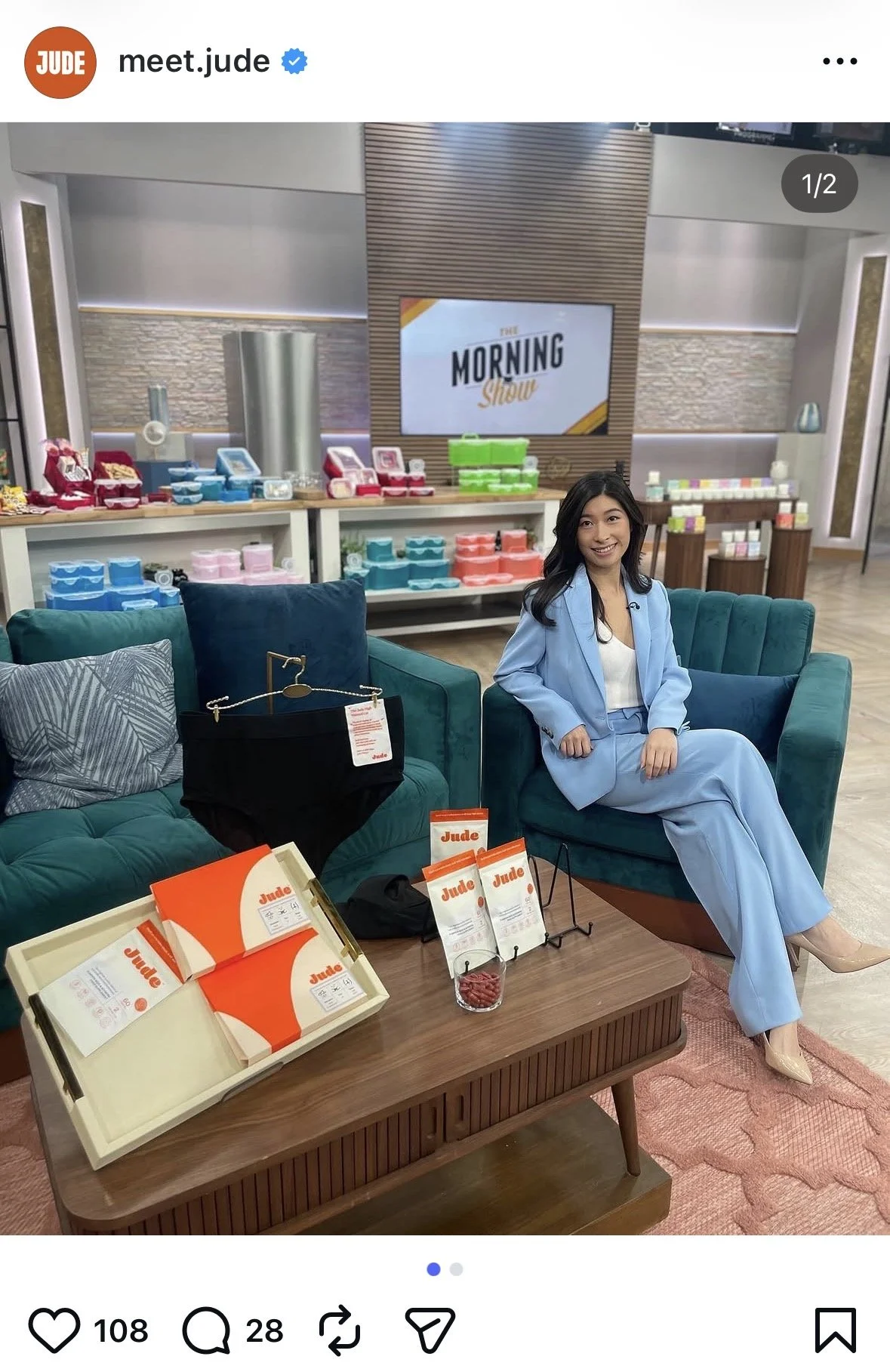 A woman sitting on a teal sofa in a TV studio, smiling, dressed in a light blue suit. In front of her is a wooden table with Jude brand product displays including orange and white boxes, pamphlets, and a small container of red candies. The background features shelves with colorful products, a large flat-screen TV with the text 'The Morning Show,' and bright studio lighting.