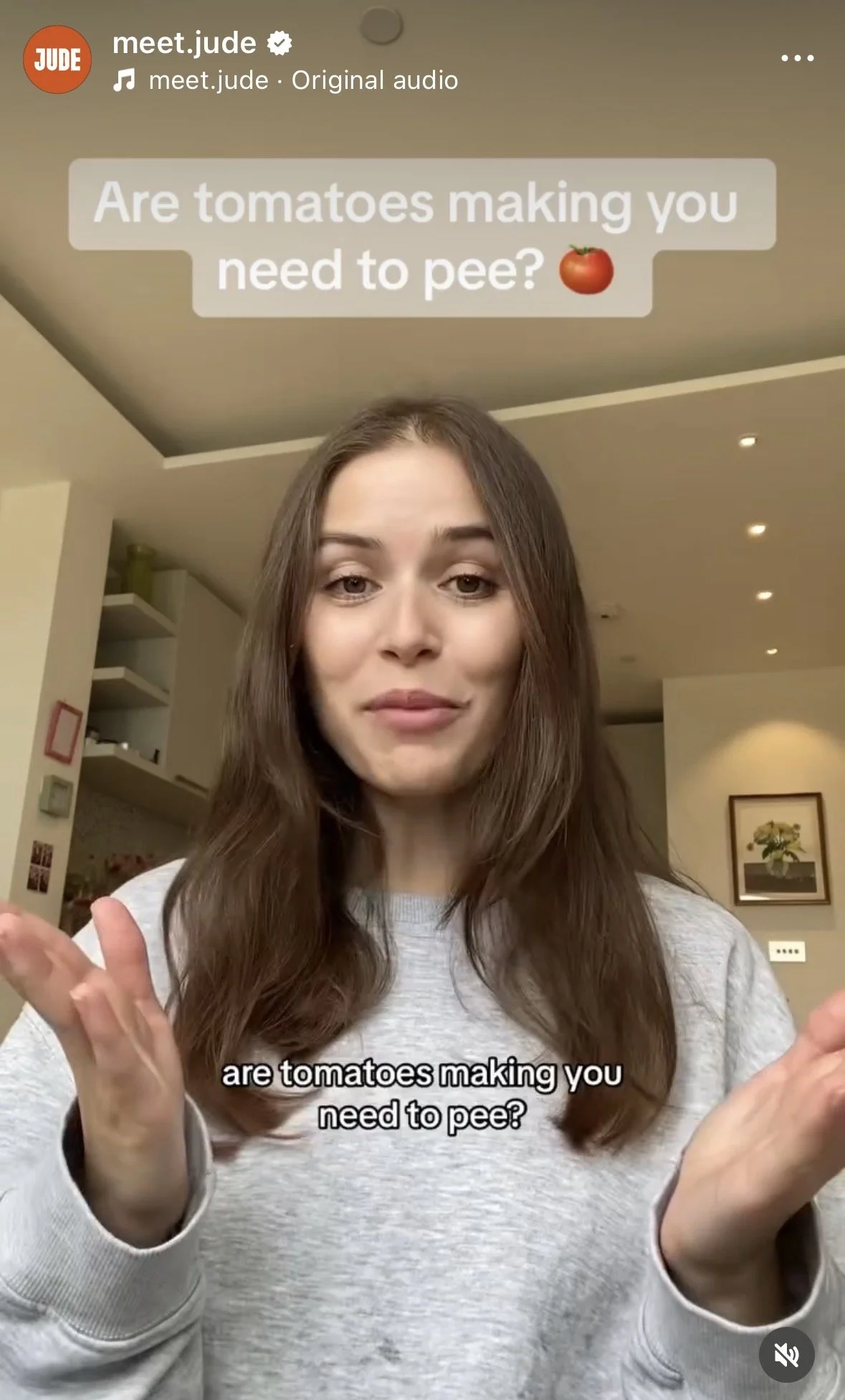 A woman with long brown hair wearing a light gray sweatshirt, gesturing with her hands, standing in a room with beige walls, shelves, and a framed picture on the wall. Text on the image asks, 'Are tomatoes making you need to pee?'.