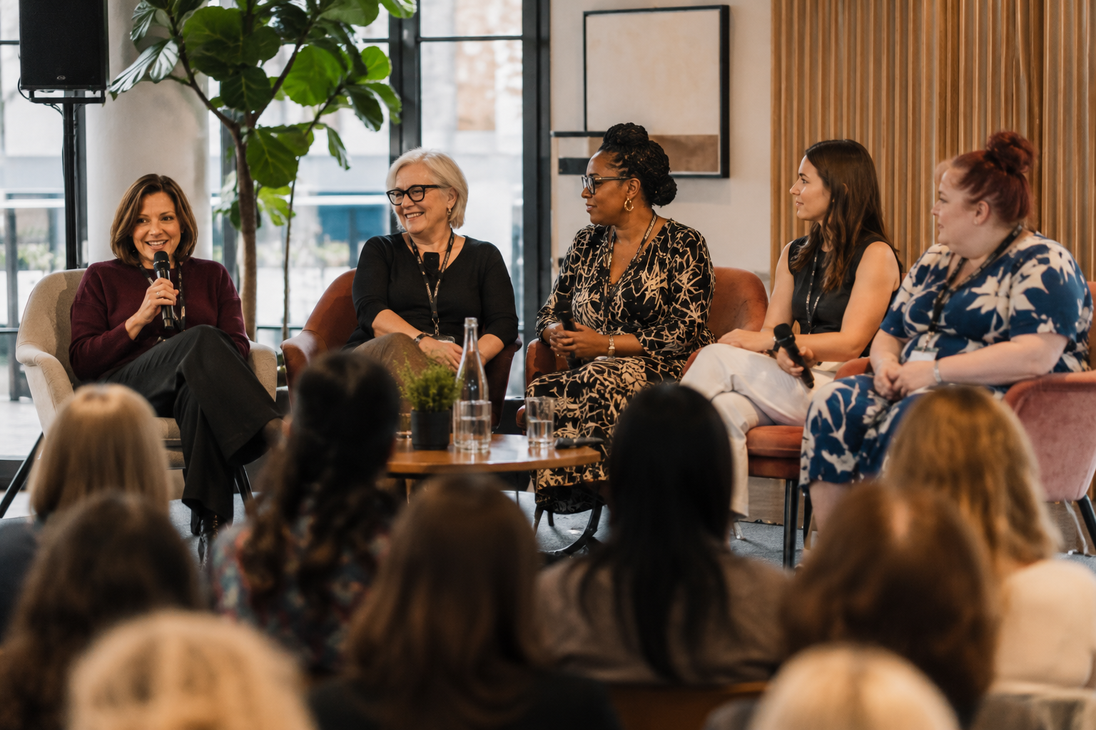 Five women seated on a panel in a modern indoor setting, with an audience in front, engaging in a discussion, some with microphones in hand.