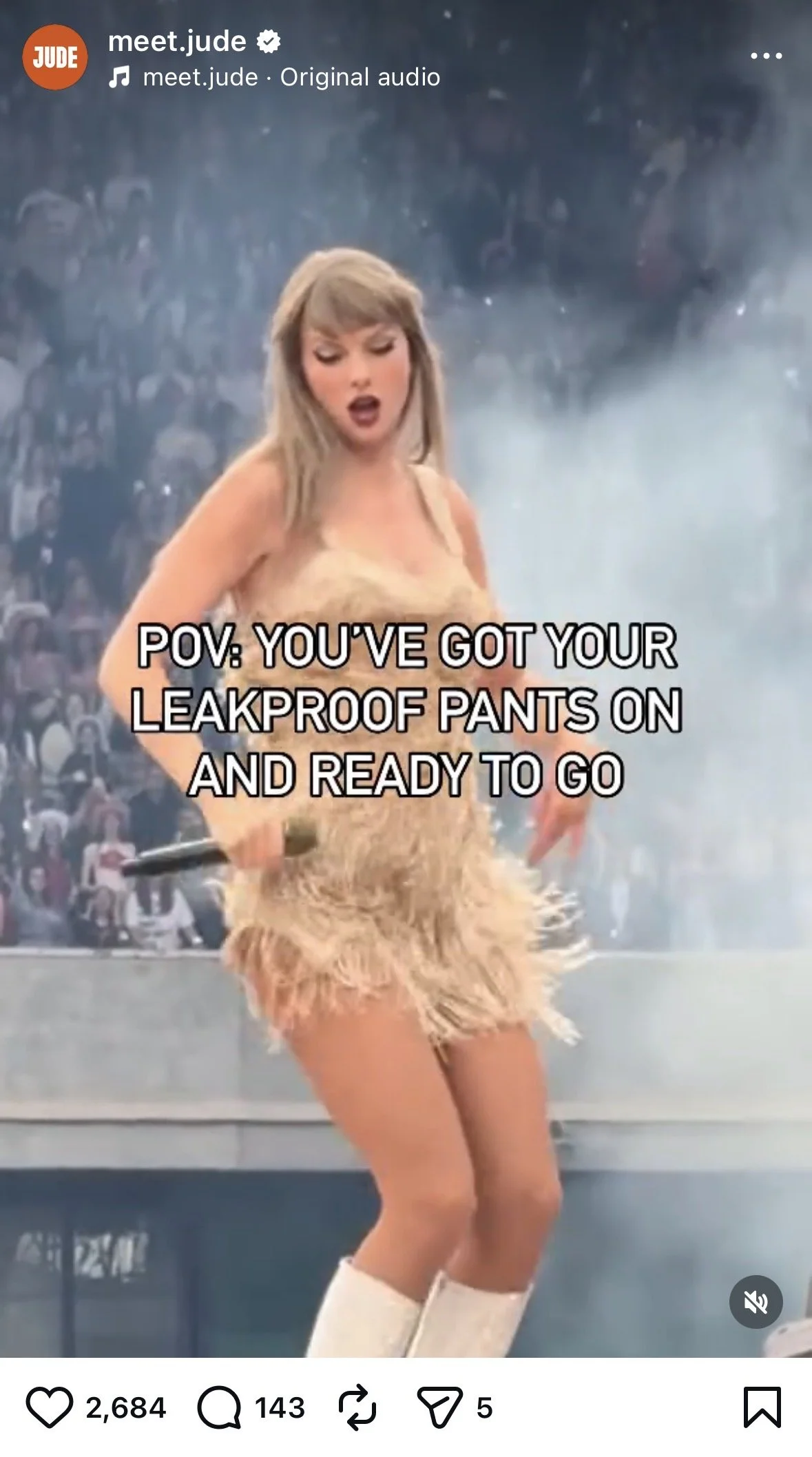 A woman performing on stage, holding a microphone, wearing a fuzzy beige outfit, white knee-high boots, and beige shorts. The background shows a large crowd and stage smoke.
