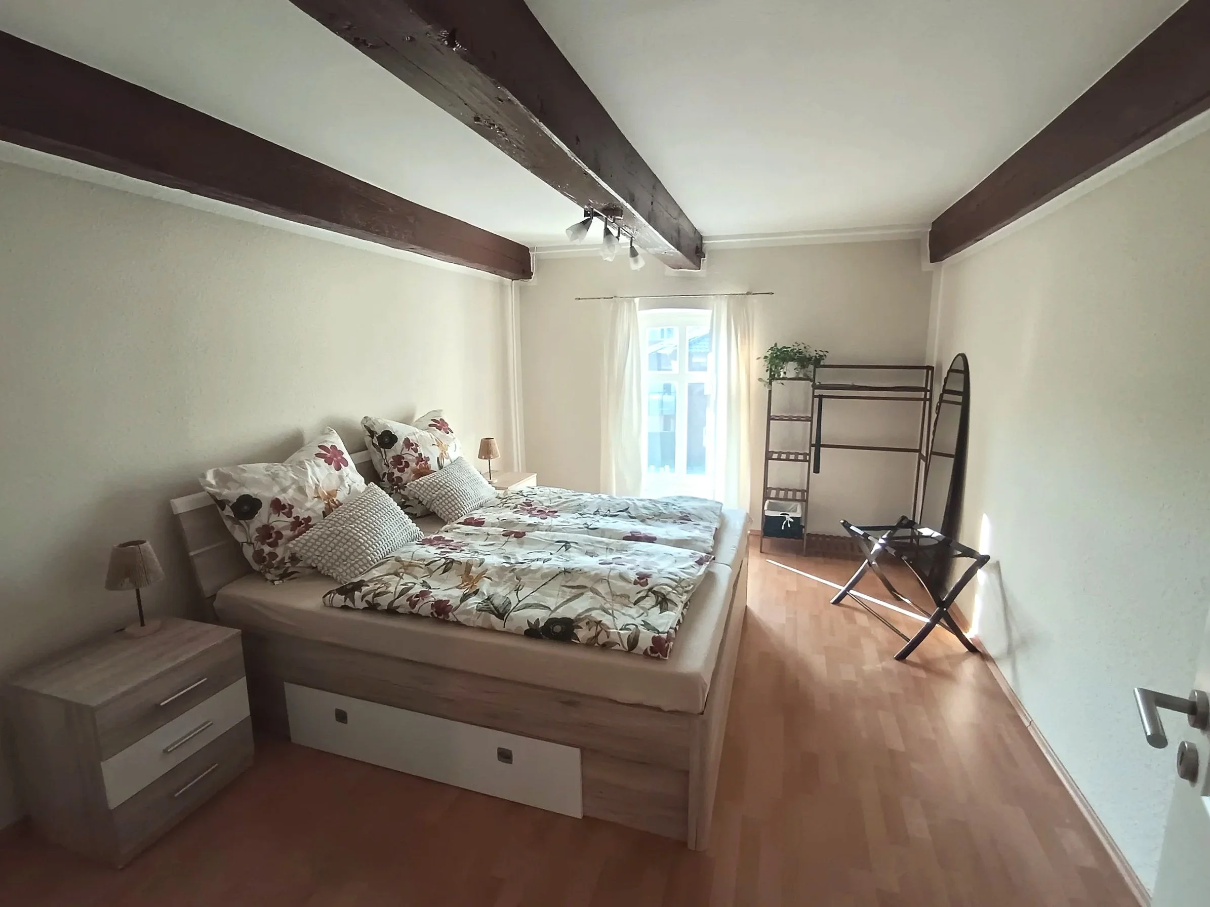 Ein Schlafzimmer mit einem Bett, Nachttischen, einer Stehlampe, einem Fenster mit weißen Vorhängen, einer Pflanze und einem Stauraum mit einem Regal, auf dem sich Gegenstände befinden. Der Raum hat Holzboden und sichtbare Holzbalken an der Decke.