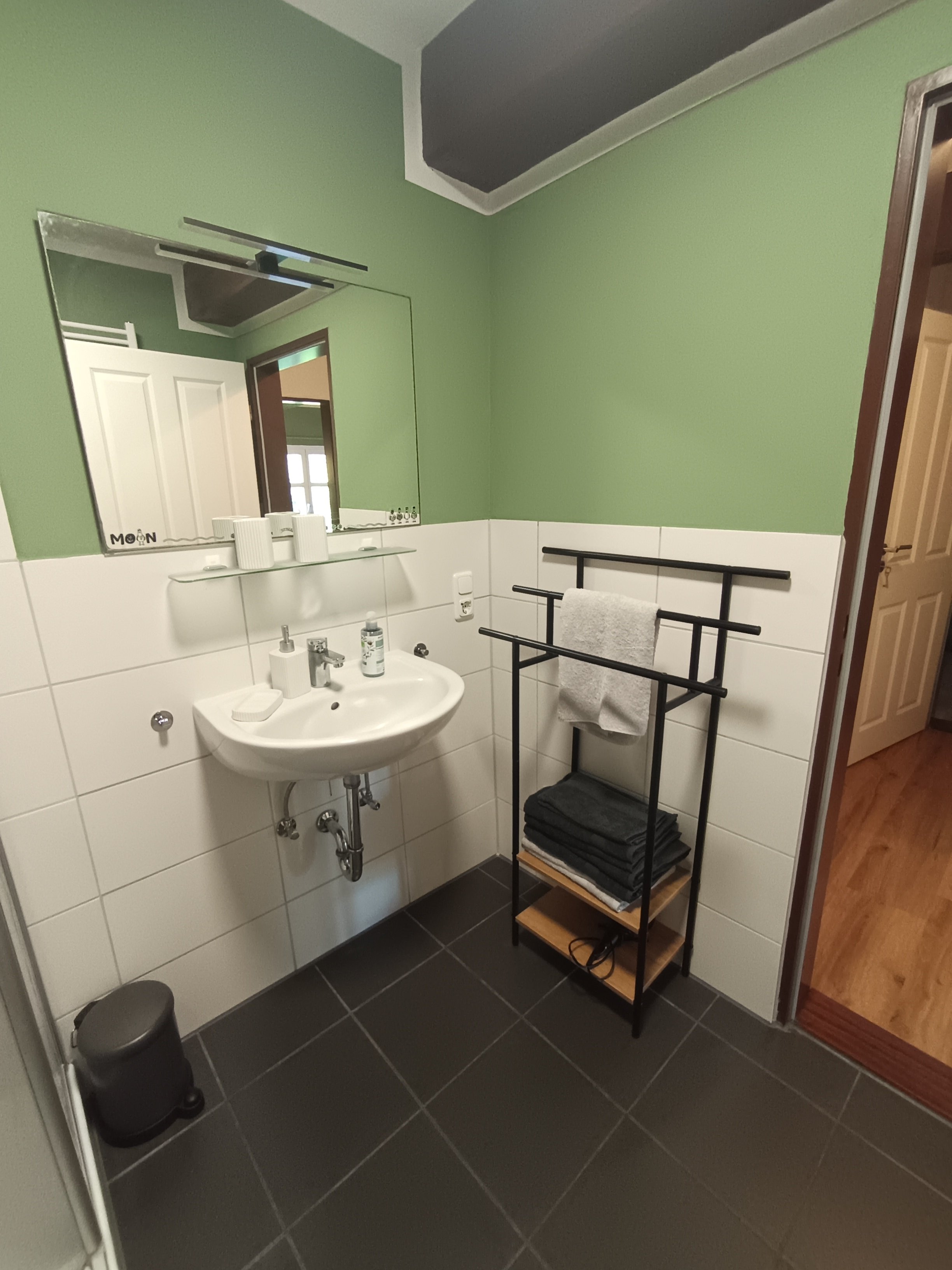 Ein Badezimmer mit einer kleinen weißen Waschtafel, einem großen Wandspiegel, einem schwarzen Handtuchständer mit einem weißen Handtuch, schwarzen Handtüchern auf einem Holregal, dunklen Fliesen auf dem Boden, und einem Holzparkett vor der Tür. Außerdem befinden sich auf der Waschbecken rechte Seite einige Pflegeprodukte und eine kleine schwarze Mülltonne auf dem Boden.