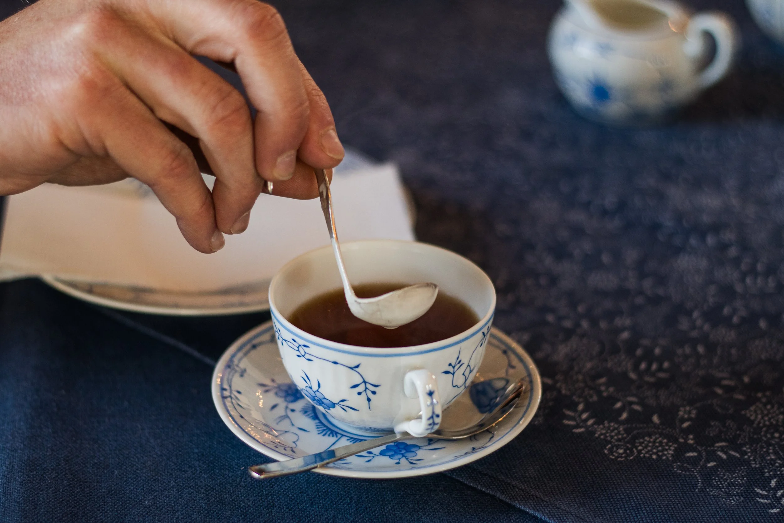 Nahaufnahme einer Hand, die Tee aus einer blauen und weißen Teetasse einschenkt, auf einem kleinen Teller mit Löffel, im Hintergrund eine weitere Tasse. Die Oberfläche ist dunkelblau.