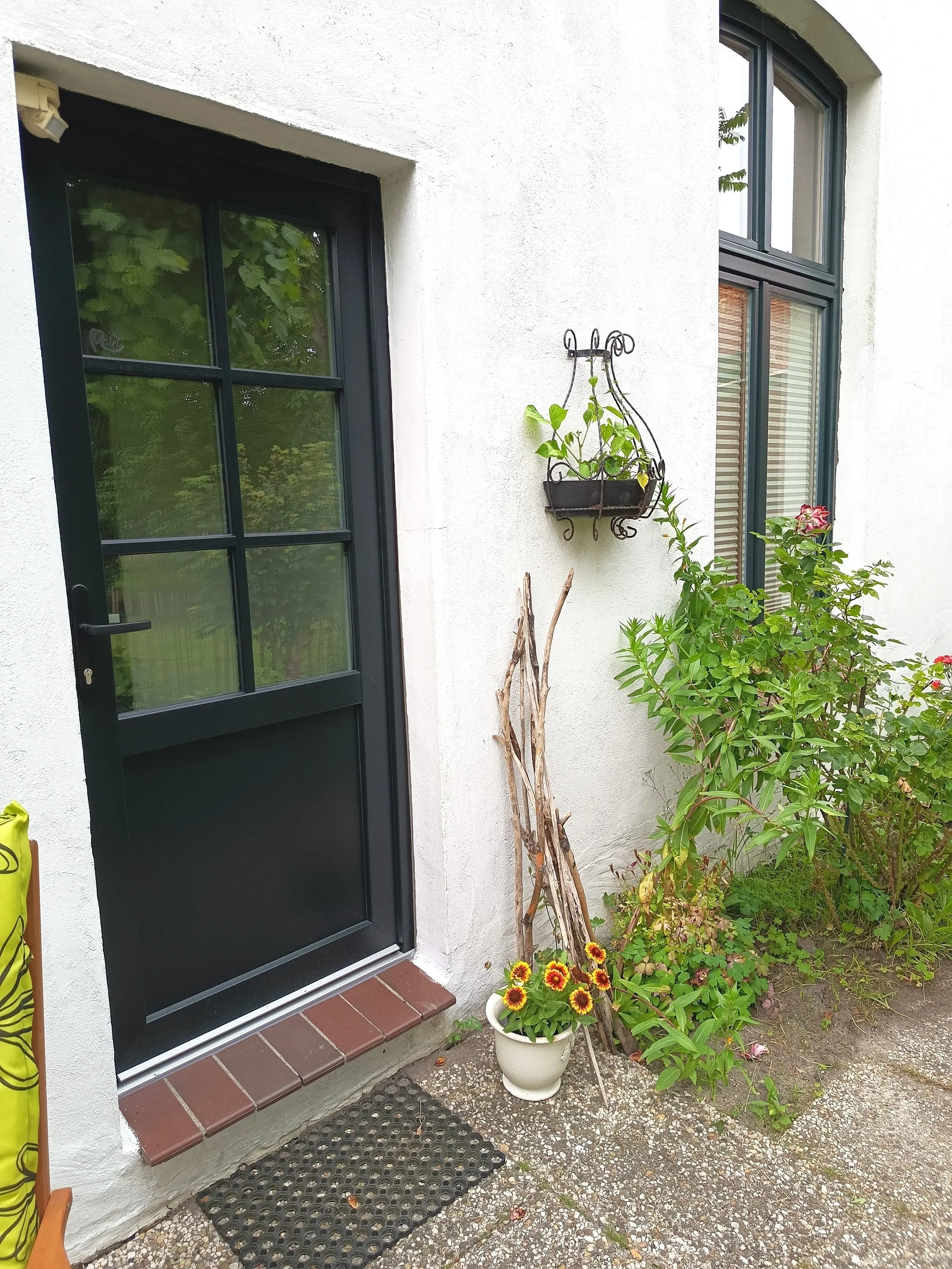 Ein Eingangsbereich eines Hauses mit einer schwarzen Tür, einer weißen Wand, einer Fenster mit grauen Rahmen, einem Blumentopf mit bunten Blumen, einigen Pflanzen und einem Holzstück. Es ist eine kleine Terrasse mit Fliesen und einem Gummimatten.