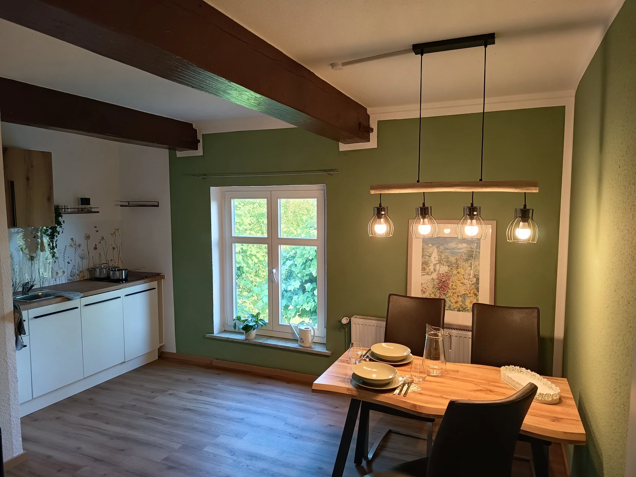 Innenaufnahme einer Küche und eines Esszimmers mit grünen Wänden, weißen Schränken, Holzdekorationen, einem Esstisch mit weißen Tellern, Weingläsern und schwarzen Stühlen. Im Hintergrund sind Fenster mit Blick nach draußen, eine Pflanze auf dem Fensterbrett und ein Kronleuchter mit vier Glühbirnen.