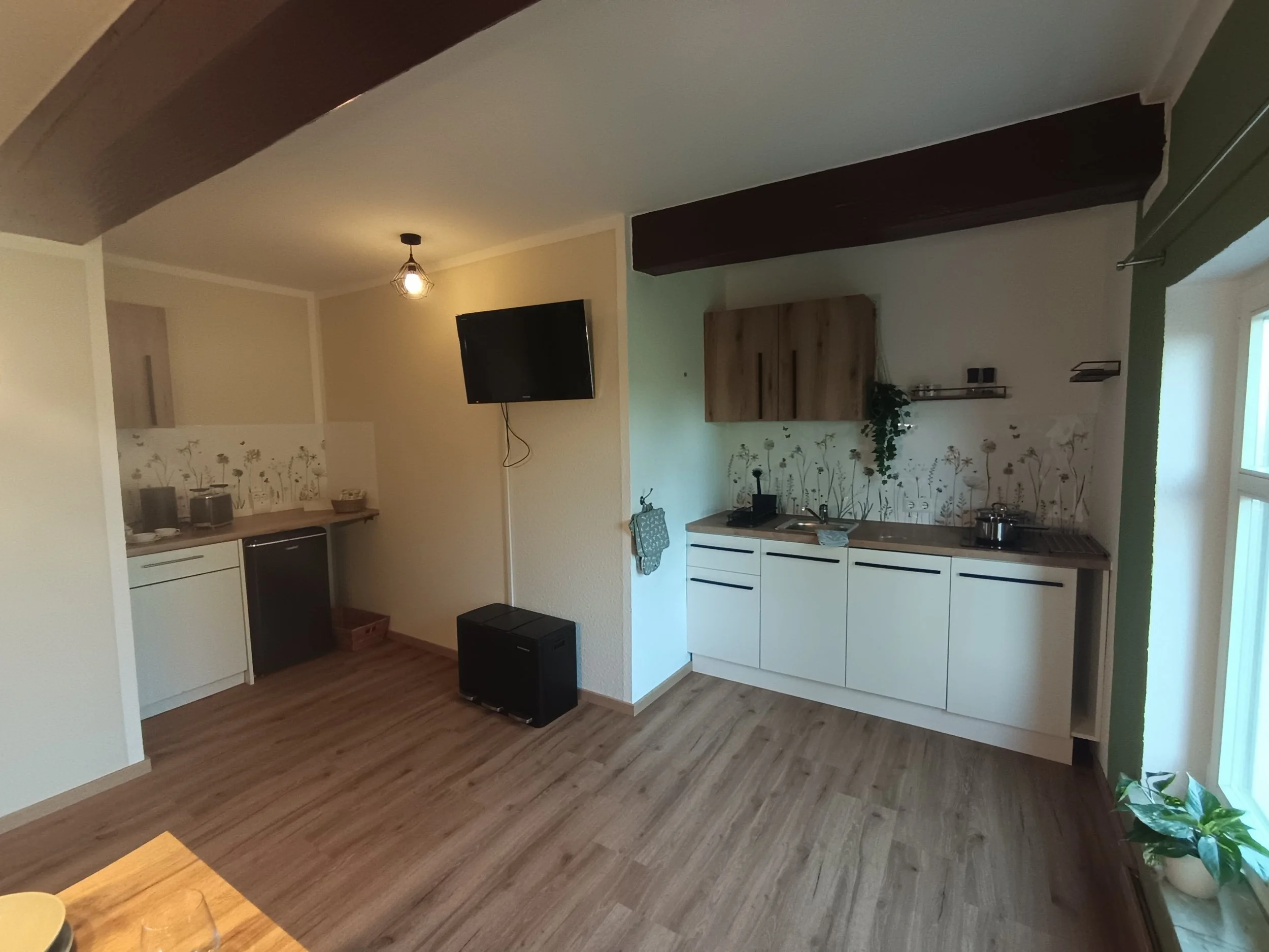 Ein Wohnzimmer mit offener Küche, Holzboden, Wanddekoration mit Blumenmotiven, Fernseher an der Wand, Küchenzeile mit weißen Schränken und Holzarbeitsplatte, grüne Wand auf der rechten Seite, Pflanzen im Raum.