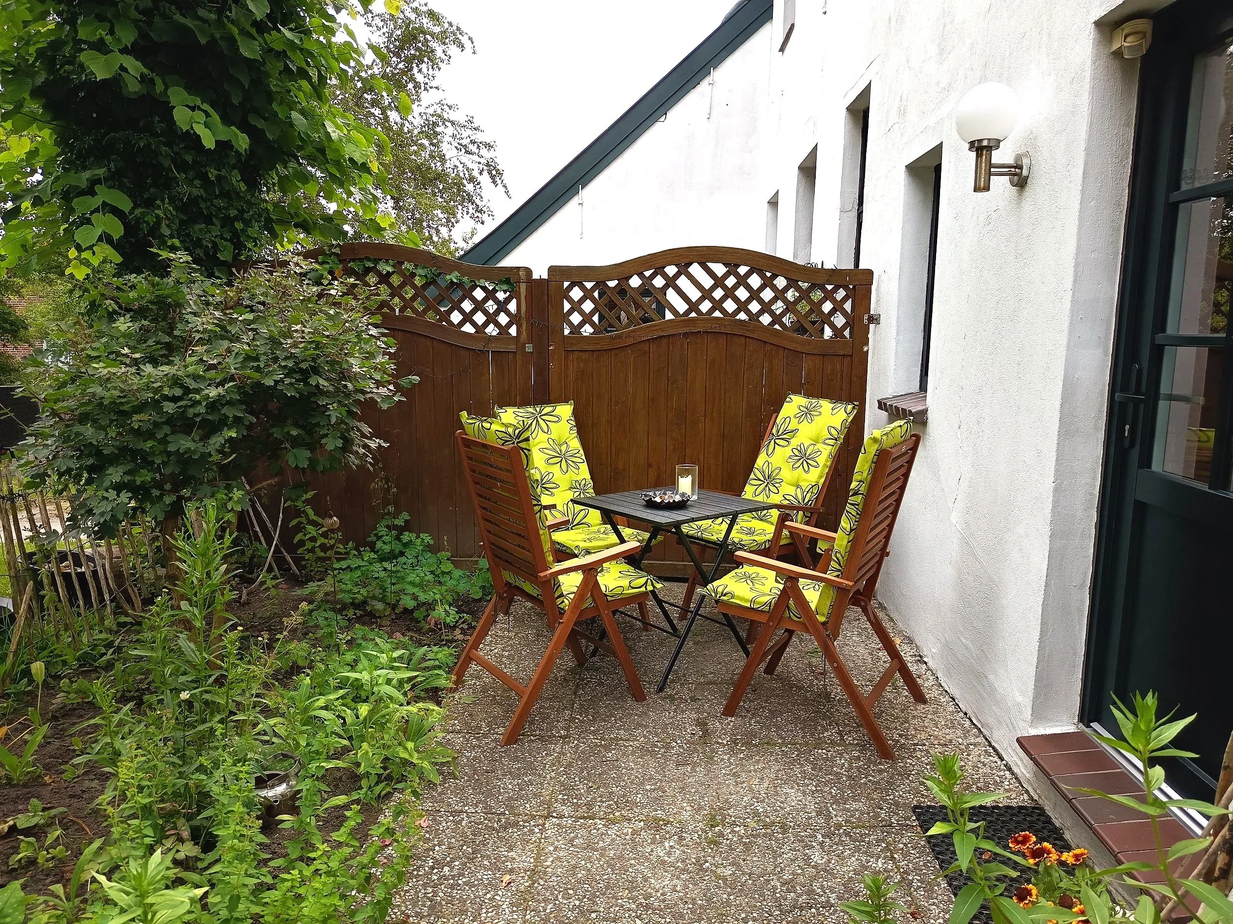 Ein Garten mit einem Holz-Tor, einem Tisch mit Sitzgelegenheiten, Pflanzen und Bäumen vor einem weißen Haus.