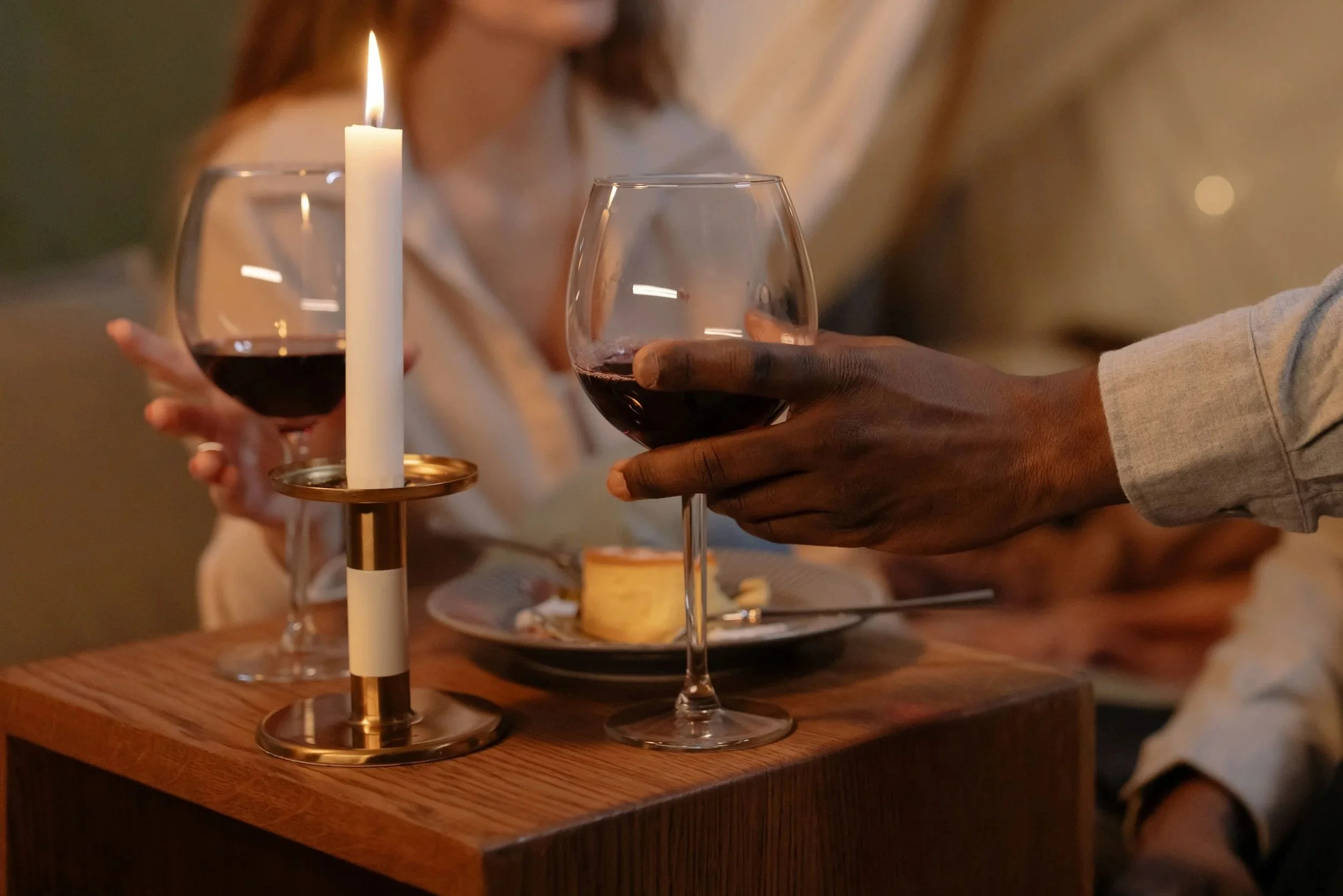 People clinking glasses of red wine in a cozy, warmly lit setting with a candle on a wooden table.