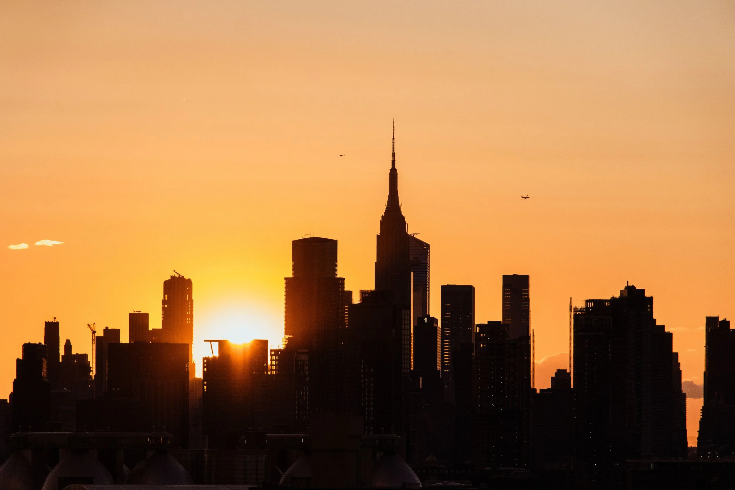 Sunset behind the New York City skyline, featuring the Empire State Building along with other tall buildings, and airplanes flying across the sky.
