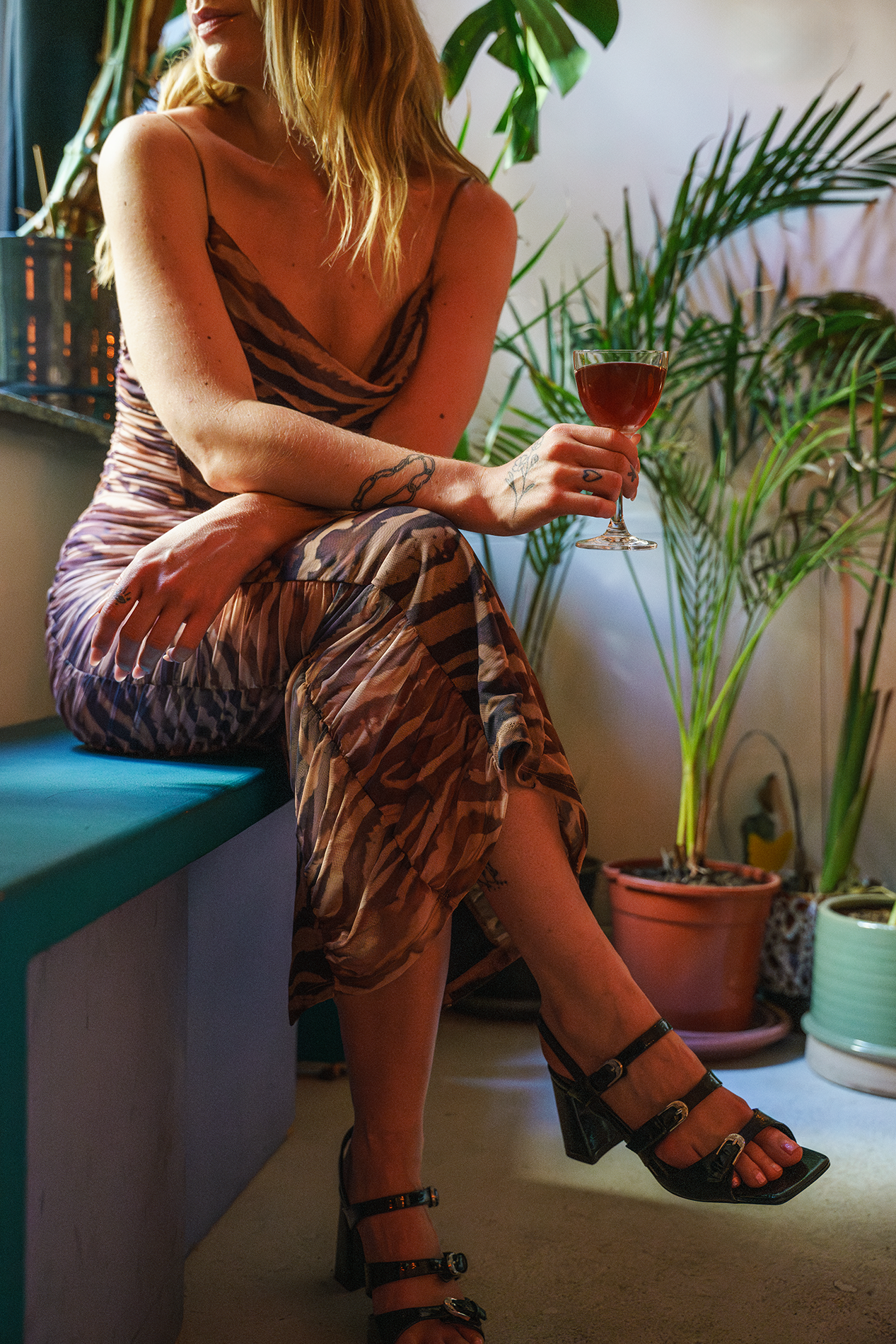 Meadow Munro with blonde hair, dressed in a zebra-print dress, is sitting on a bench in a room filled with potted plants. She is holding a glass of red wine, and her face is partially visible.