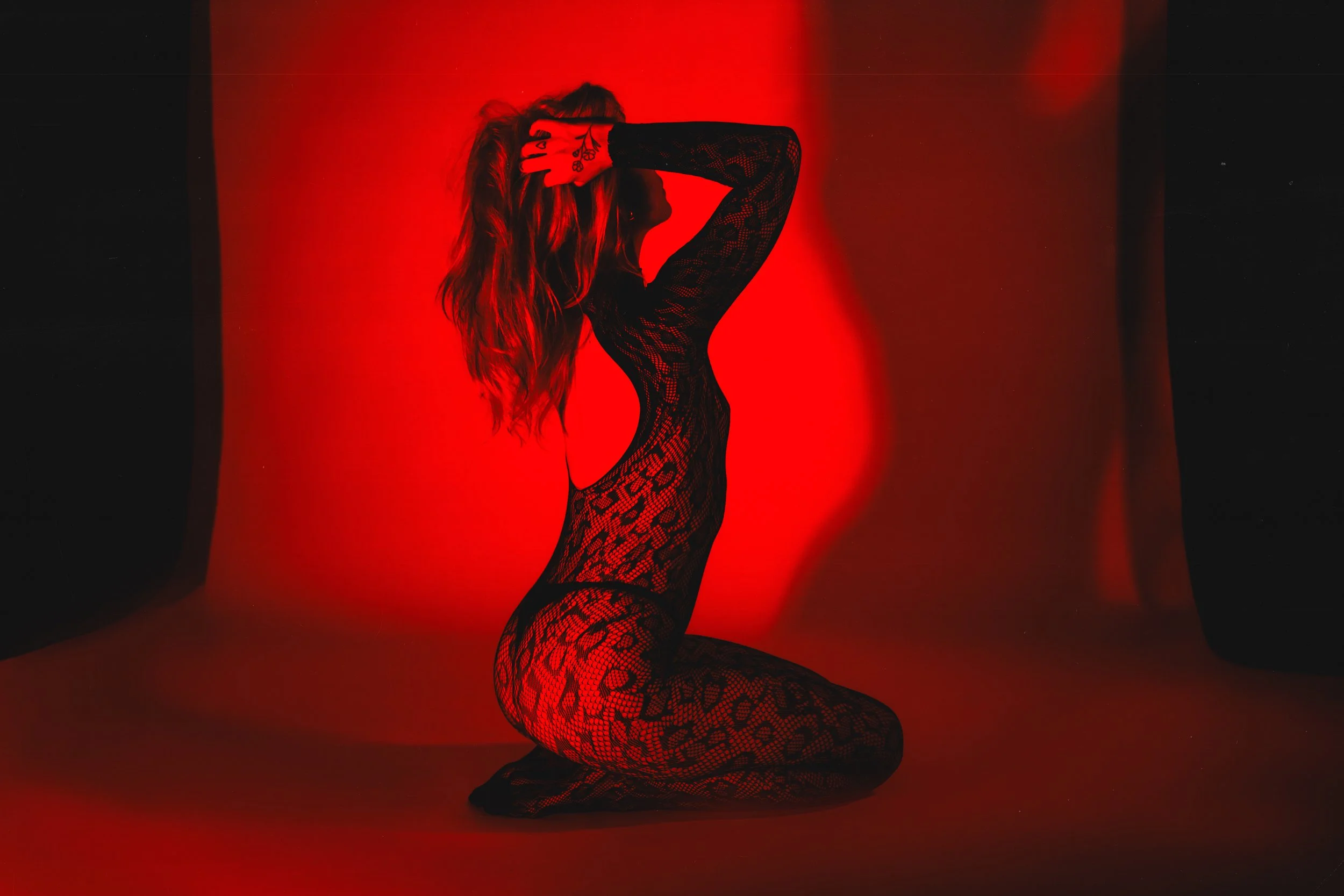 Meadow Munro with long hair, wearing a black lace bodysuit, is posing with one hand on their head. The background is a gradient of red and black.