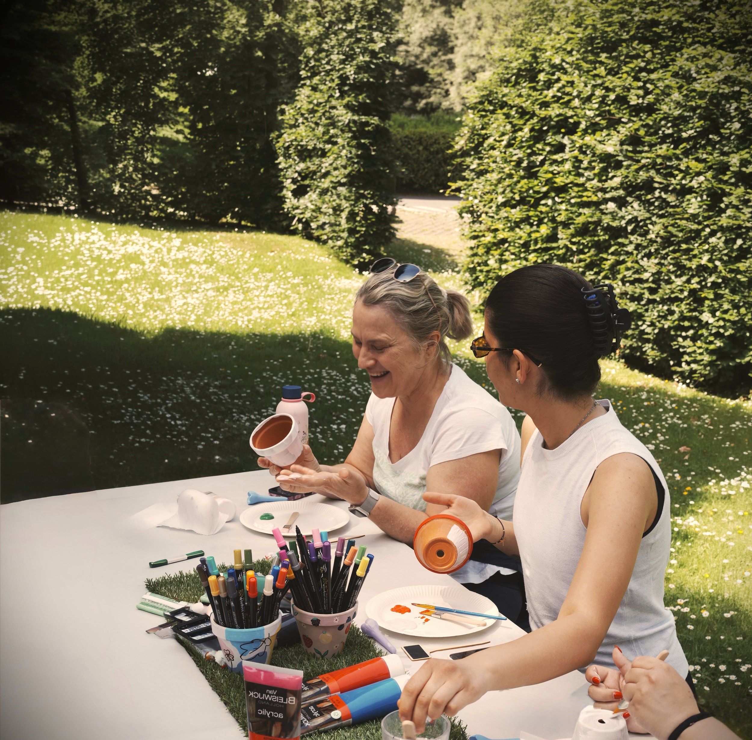 Two women laughing while painting terracotta pots at an outdoor art workshop during a creative retreat