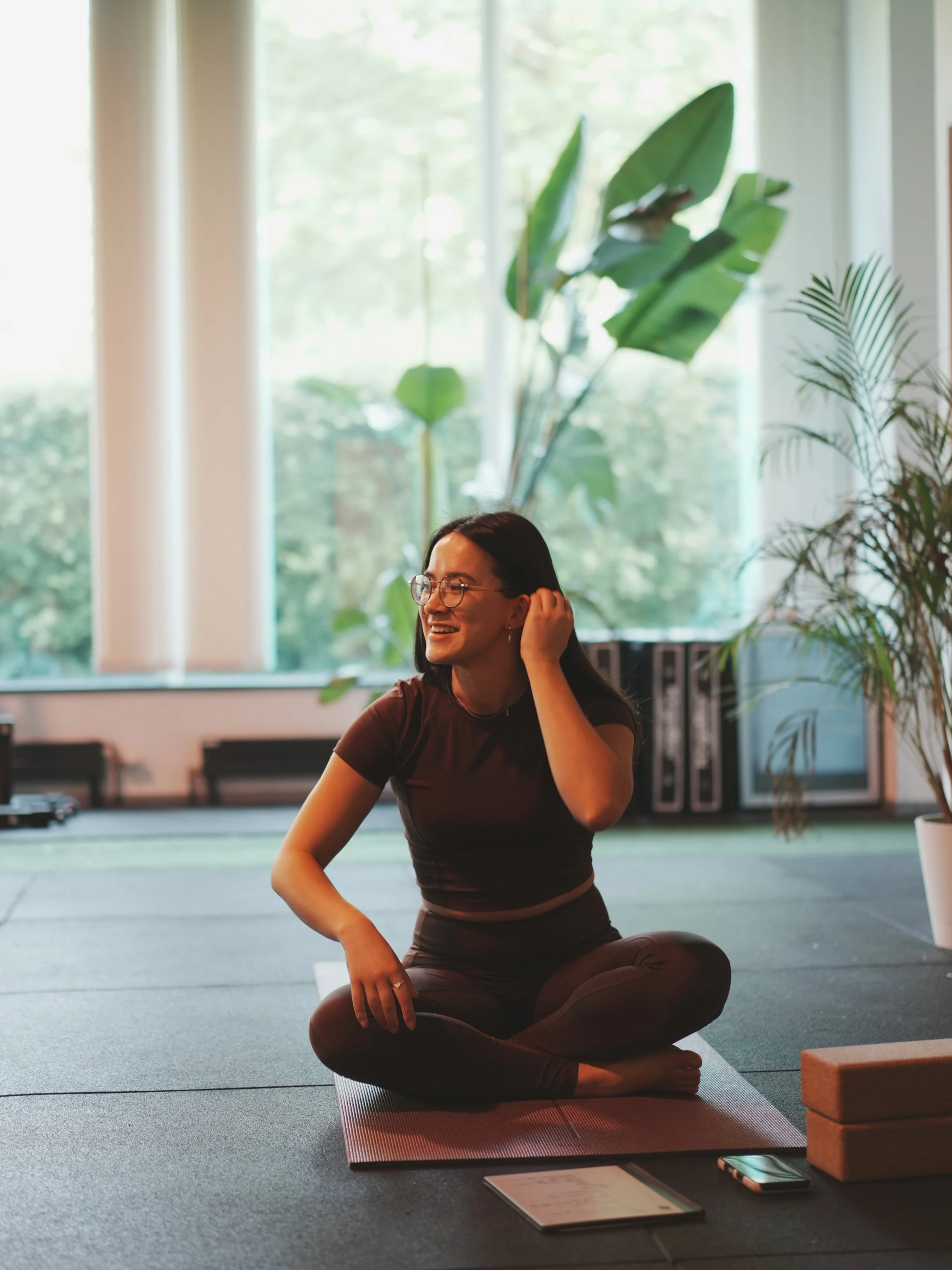 A woman sitting cross-legged on a yoga mat in a bright room, smiling while adjusting her hair. Large windows with greenery visible outside, and plants in the background. Yoga props are on the floor nearby.