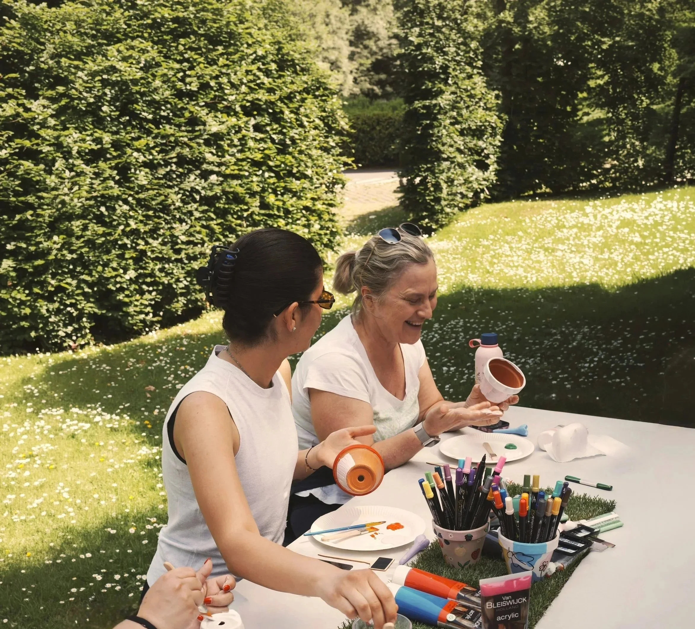 People sitting at a table outdoors, engaging in arts and crafts with paints and markers in a garden setting.
