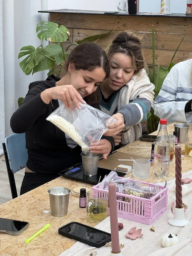 Two women smiling while measuring out white wax pellets for candle making during a creative workshop at a wellness retreat.
