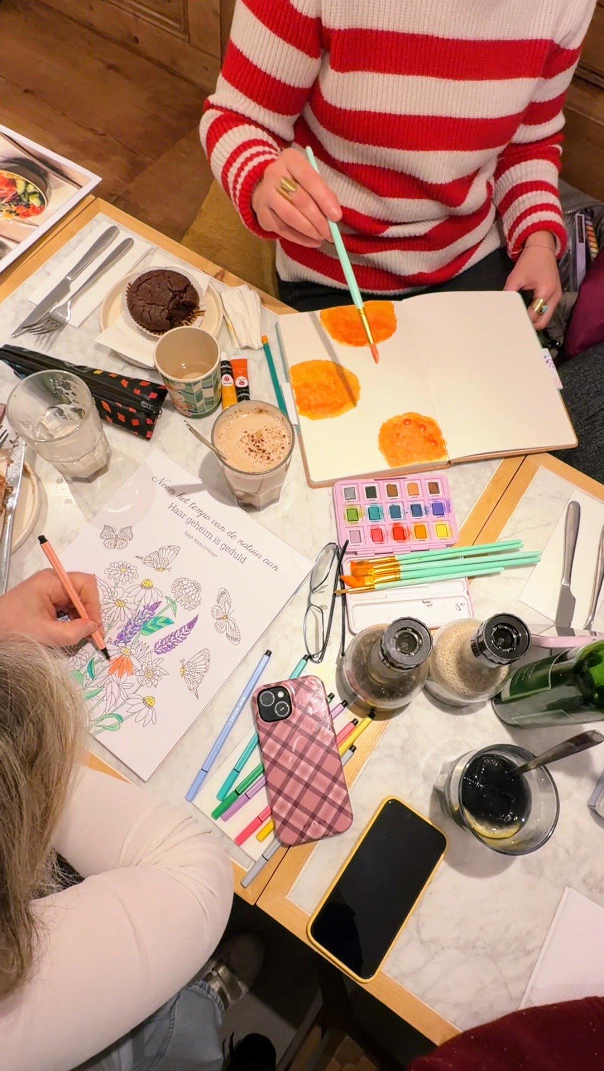 Two women painting watercolors and coloring at a cafe table with coffee and pastries during a creative wellness retreat.