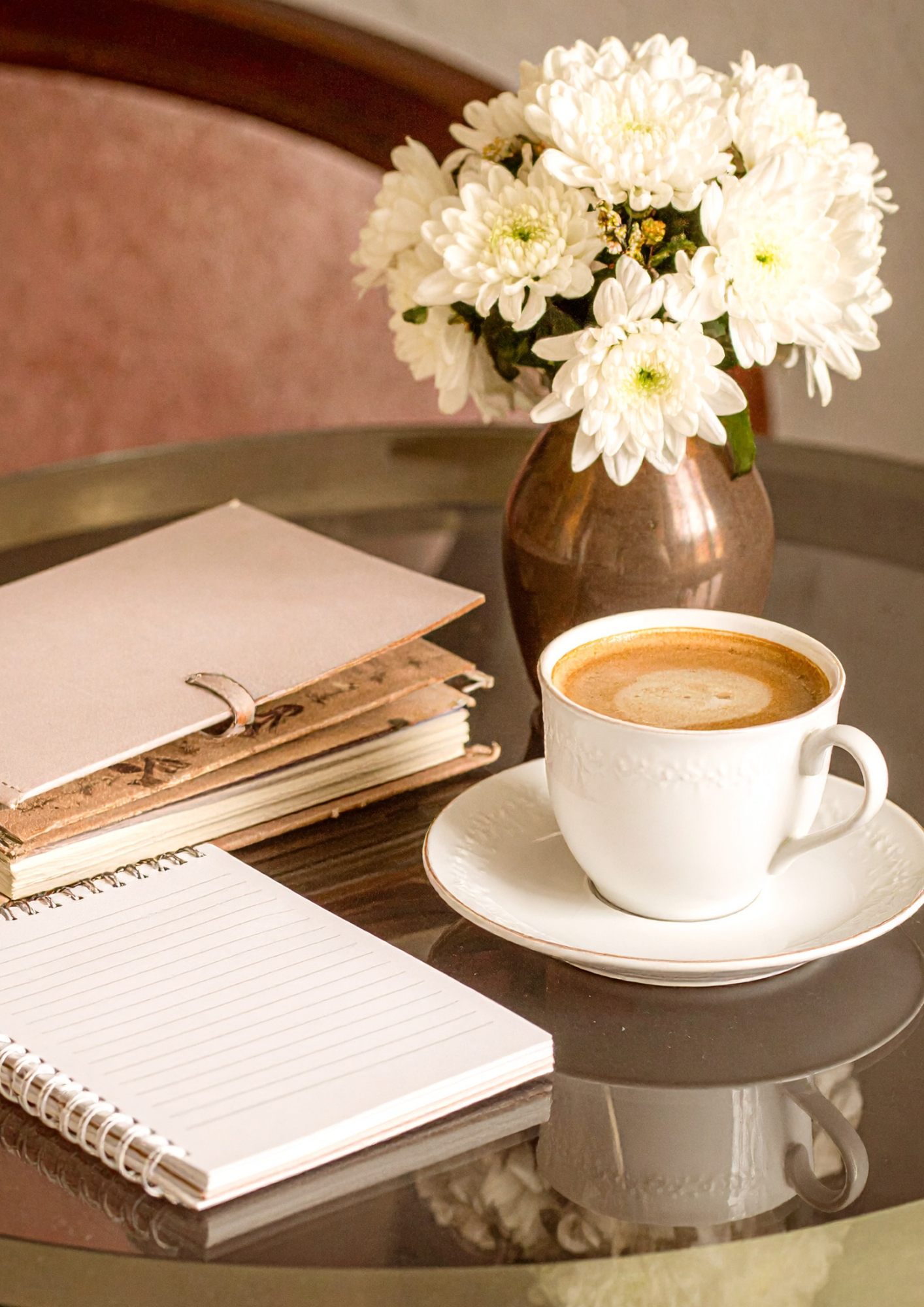 A coffee cup filled with coffee, a notebook with a ribbon marker, a plant in a brown vase with white flowers, and a lined notepad on a dark table.