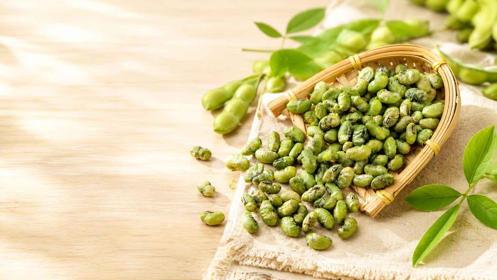Fresh green edamame beans spilling from a bamboo basket onto a beige cloth, with green leaves and a light wooden surface in the background.
