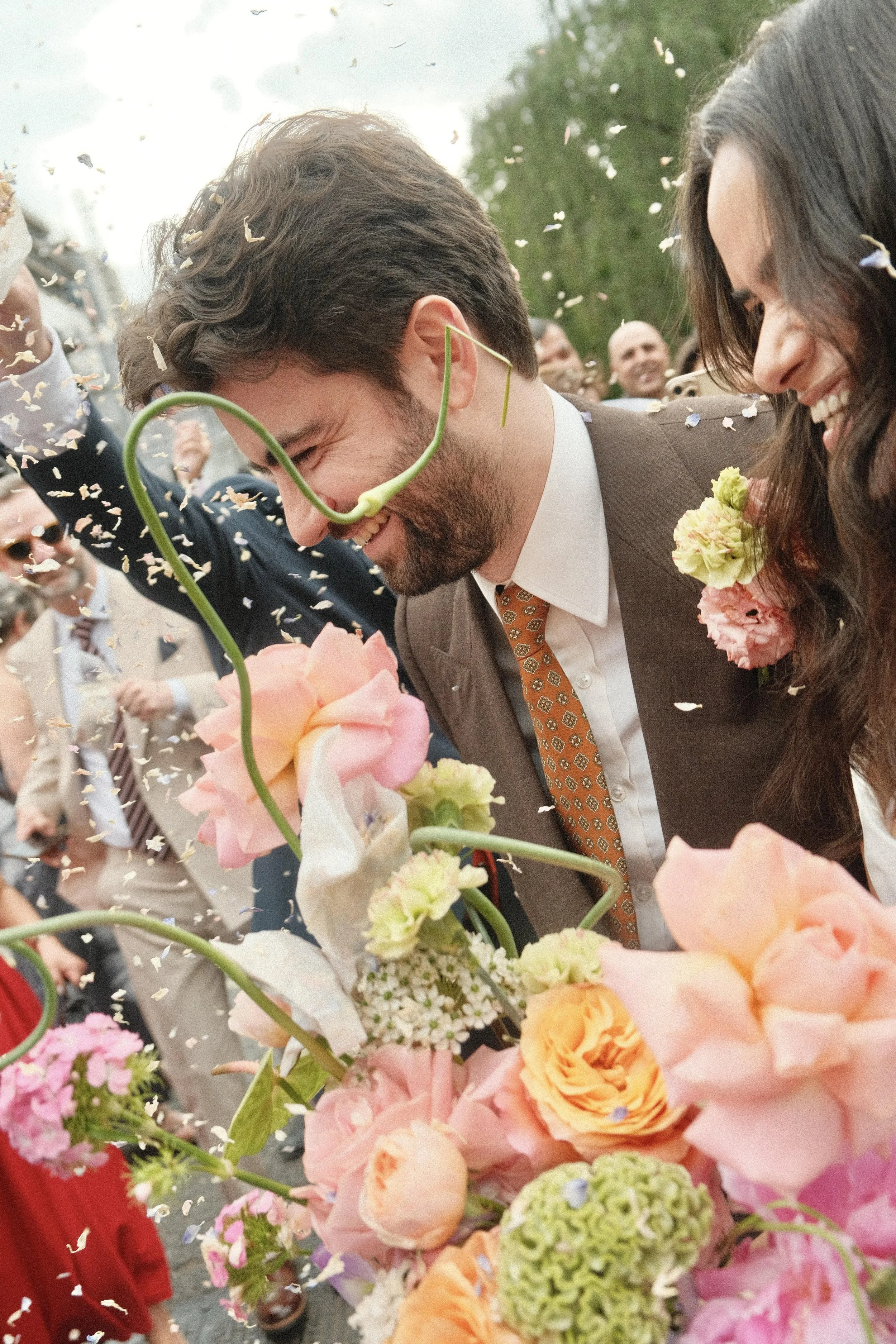 A man and woman smiling and leaning close to each other at a celebration, surrounded by colorful flowers and falling confetti, outdoors with trees and a crowd of people in the background.