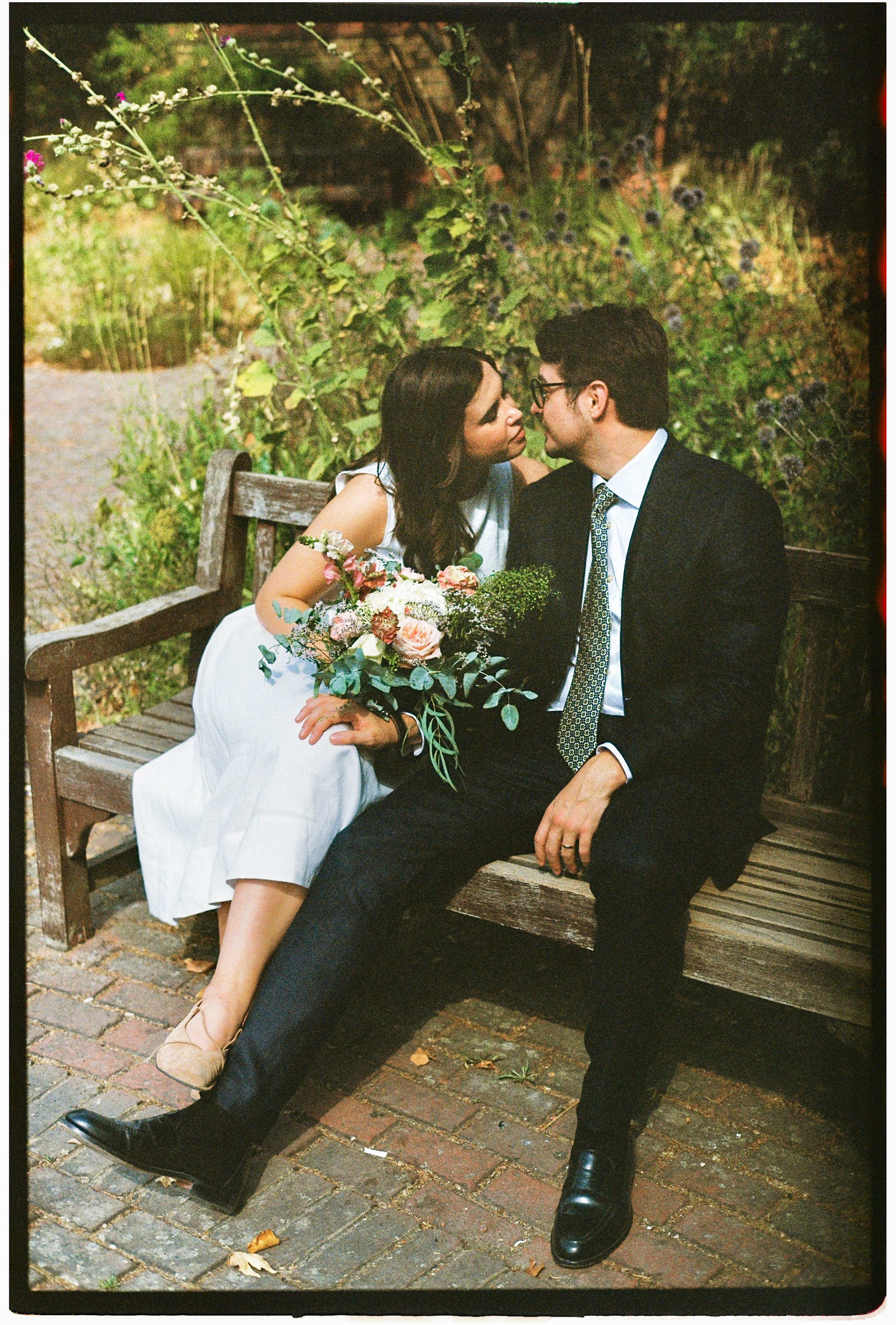 A man and woman sitting close together in Battersea Park on a wooden bench in a garden, with the woman holding a bouquet of flowers. They are leaning in as if about to kiss, surrounded by green plants and flowers.