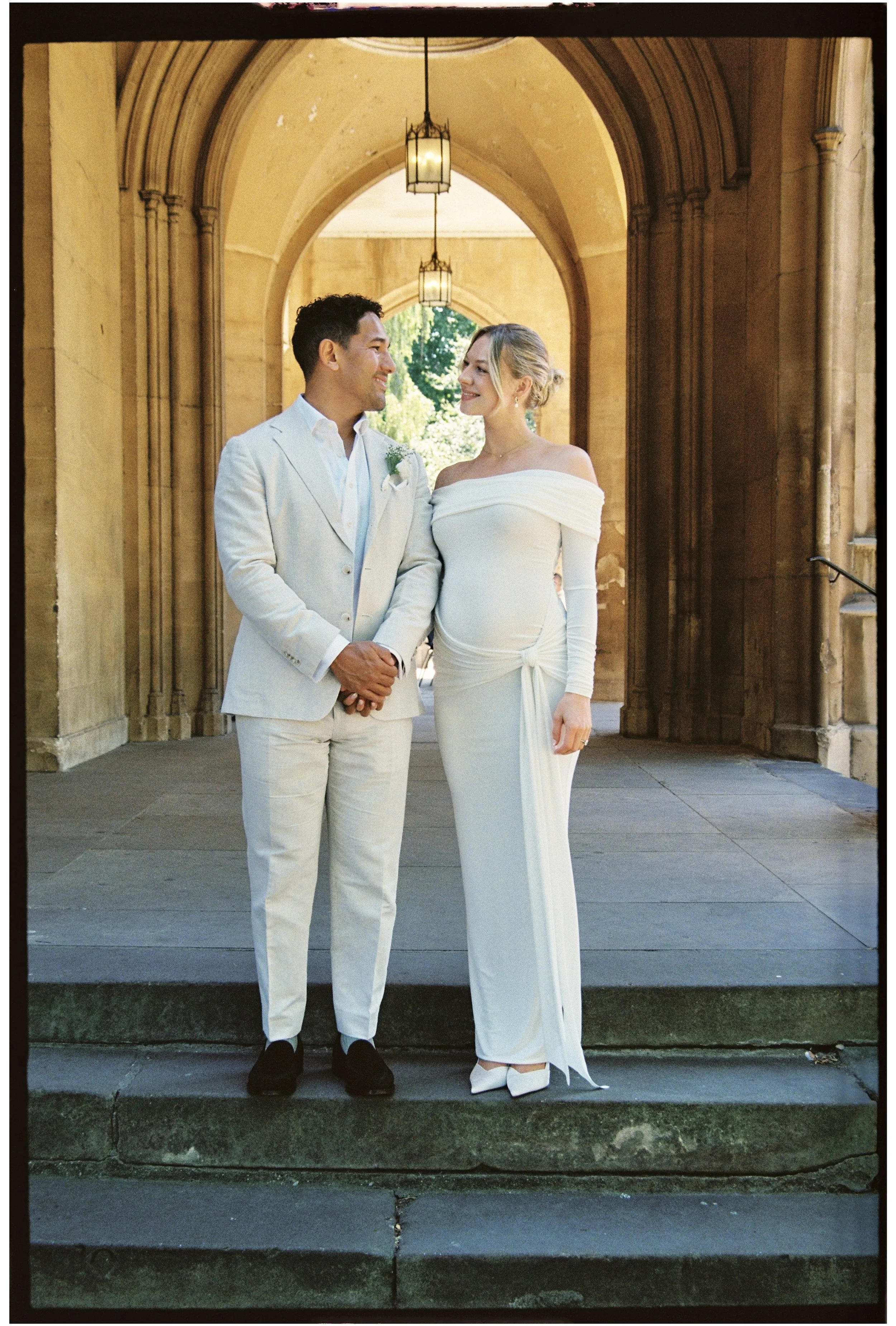 A couple dressed in wedding attire stands under an arched stone walkway with hanging lanterns, looking at each other and smiling after their ceremony at Chelsea Old Town Hall.