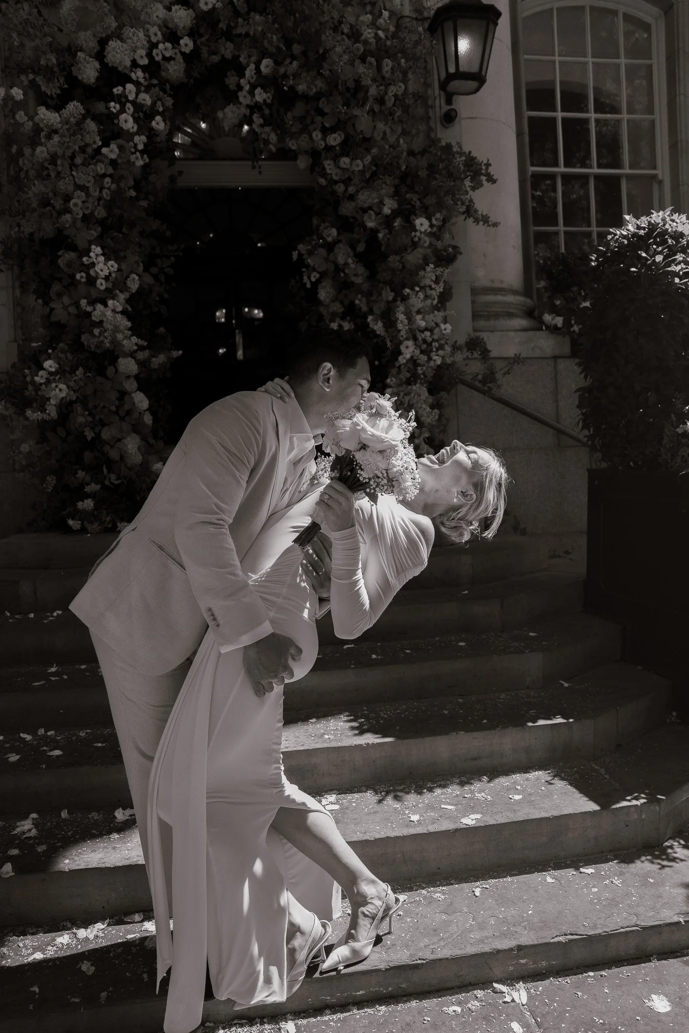 A couple dressed in wedding attire sharing a romantic dance on steps outside Old Chelsea Town Hall with floral decorations and a window.