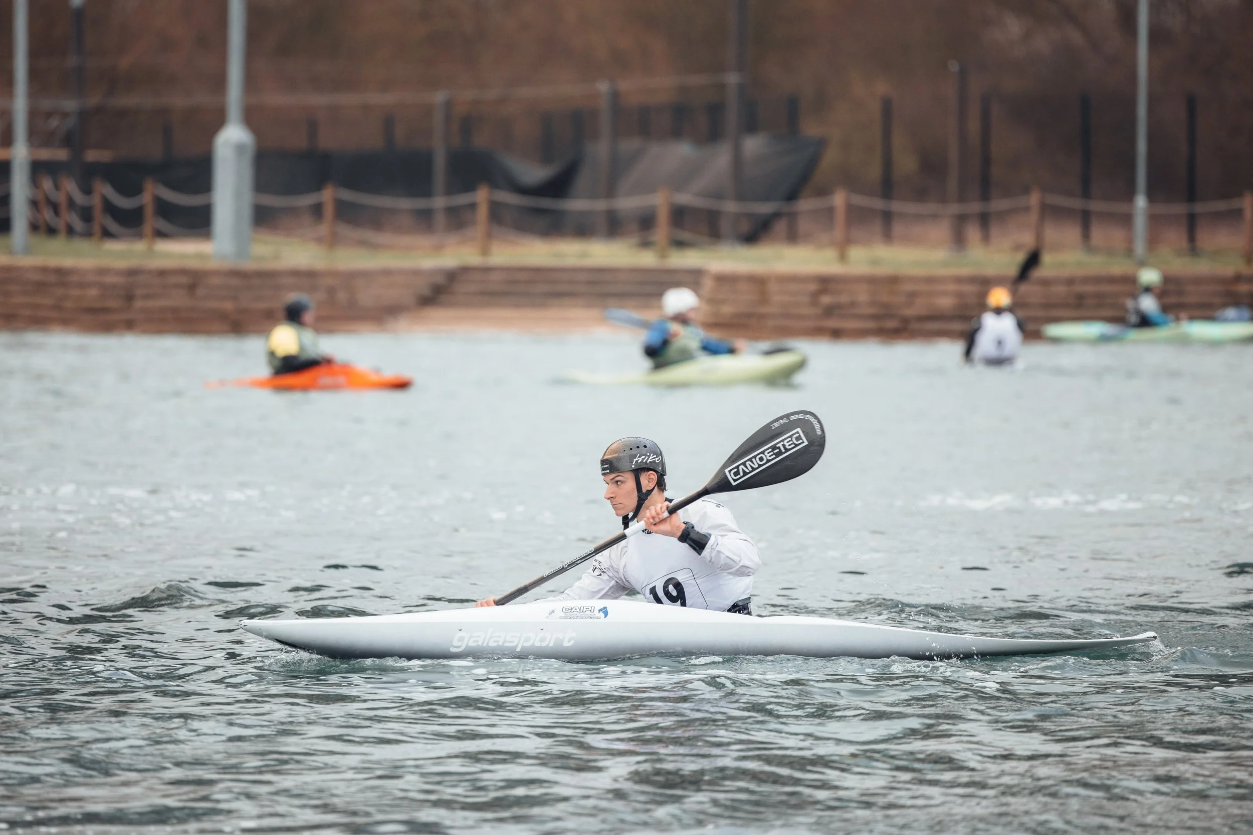 A person paddling a white kayak with the number 19 on it on a body of water, with other kayakers in the background and a wooden dock on the shore.