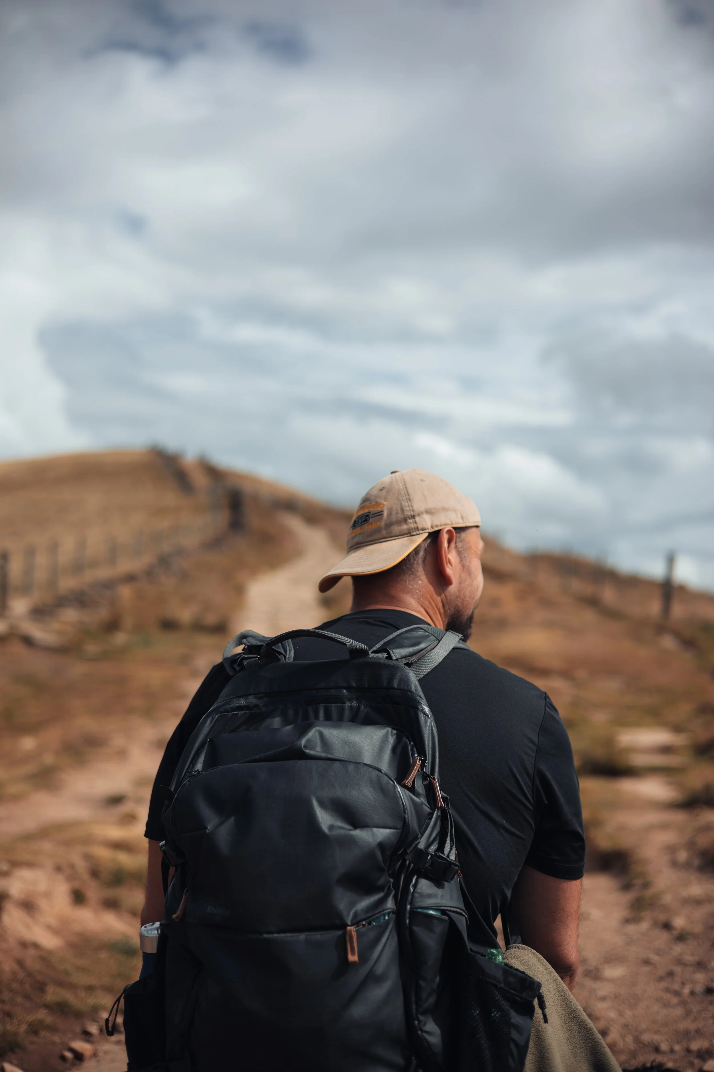 Person with a backpack and hat walking on a dirt trail in a hilly, mountainous landscape under cloudy sky.